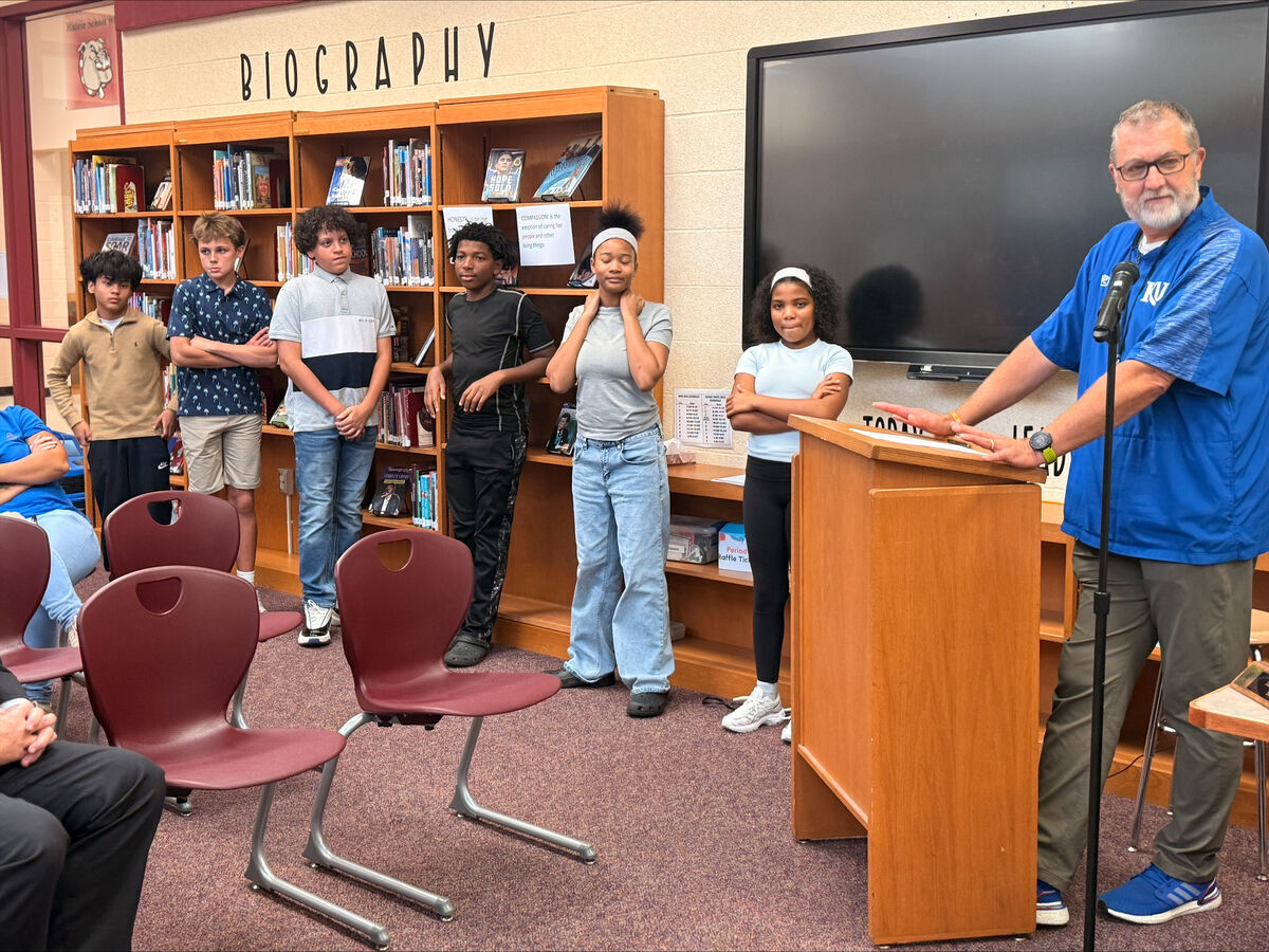 A teacher stands at a podium in a school library, addressing a group of students standing in front of bookshelves labeled 'BIOGRAPHY'.