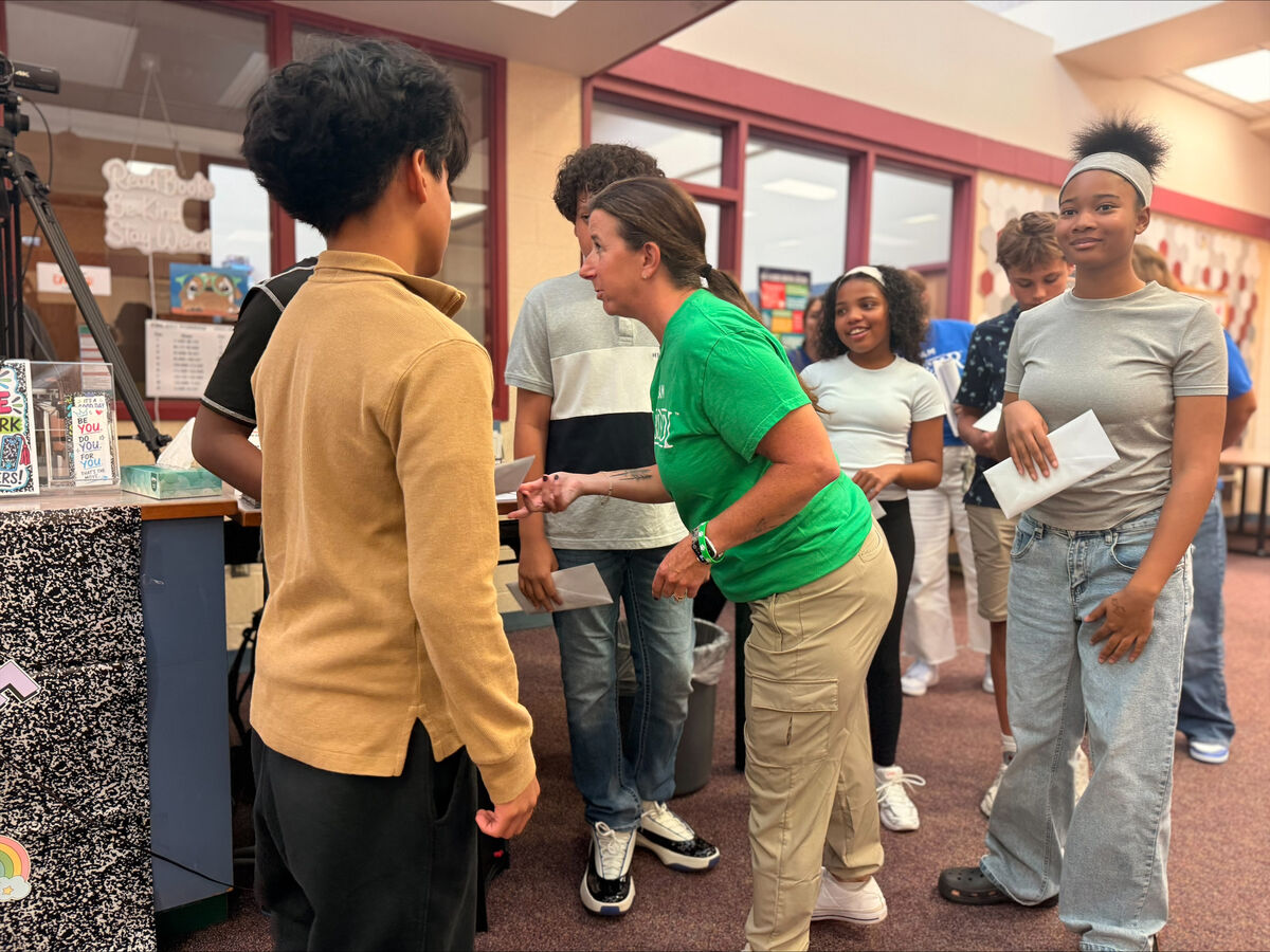 A teacher in a bright green shirt interacts with students in a brightly lit school hallway.