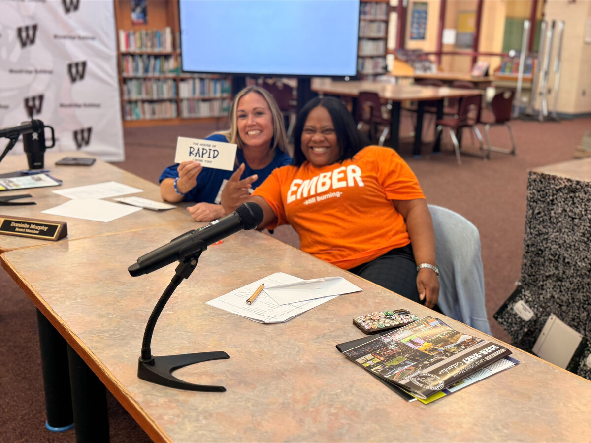 Two smiling women sit behind a large conference table in a library setting, with a microphone in the foreground.