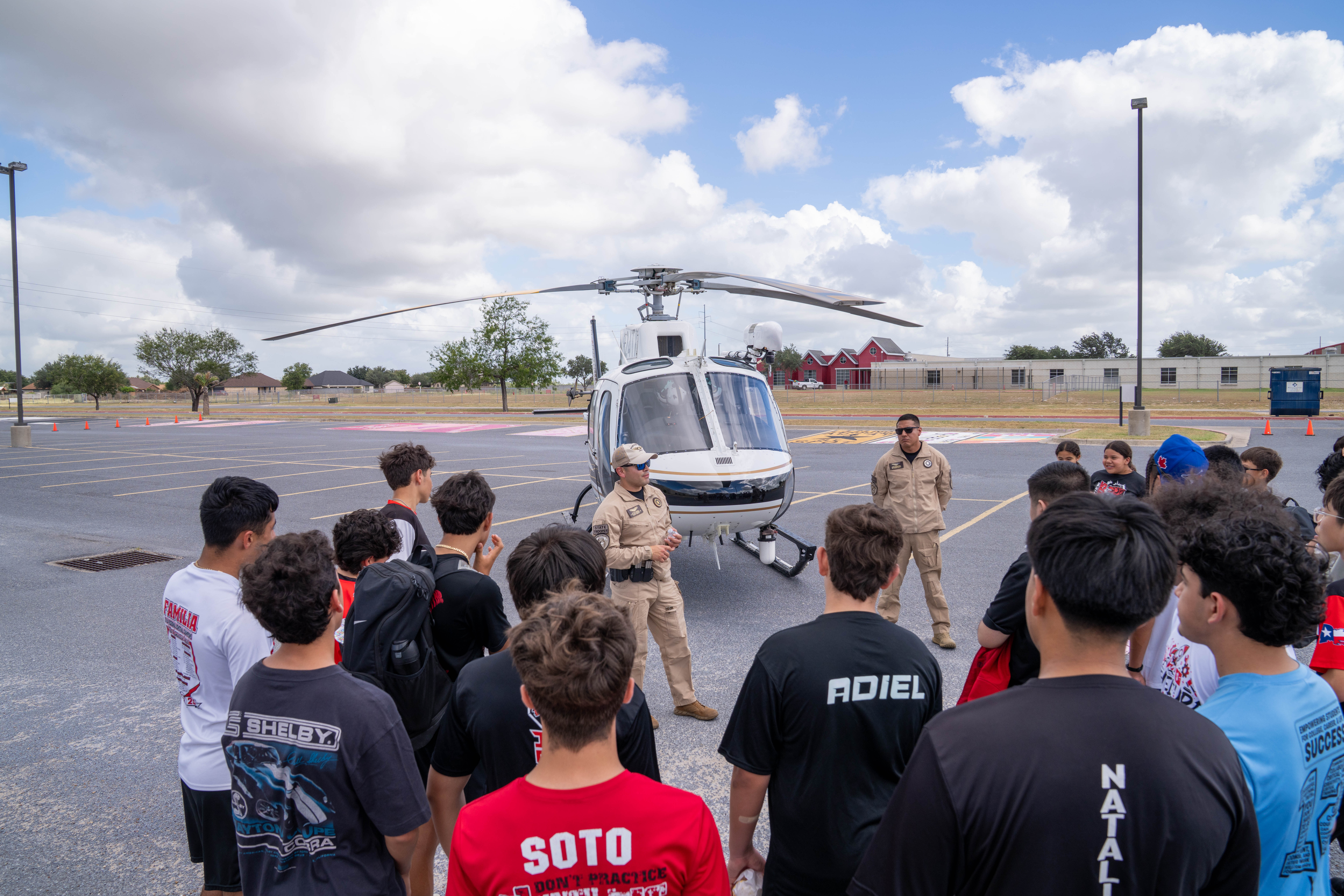 A group of students gathers around a white helicopter with two uniformed individuals standing nearby.