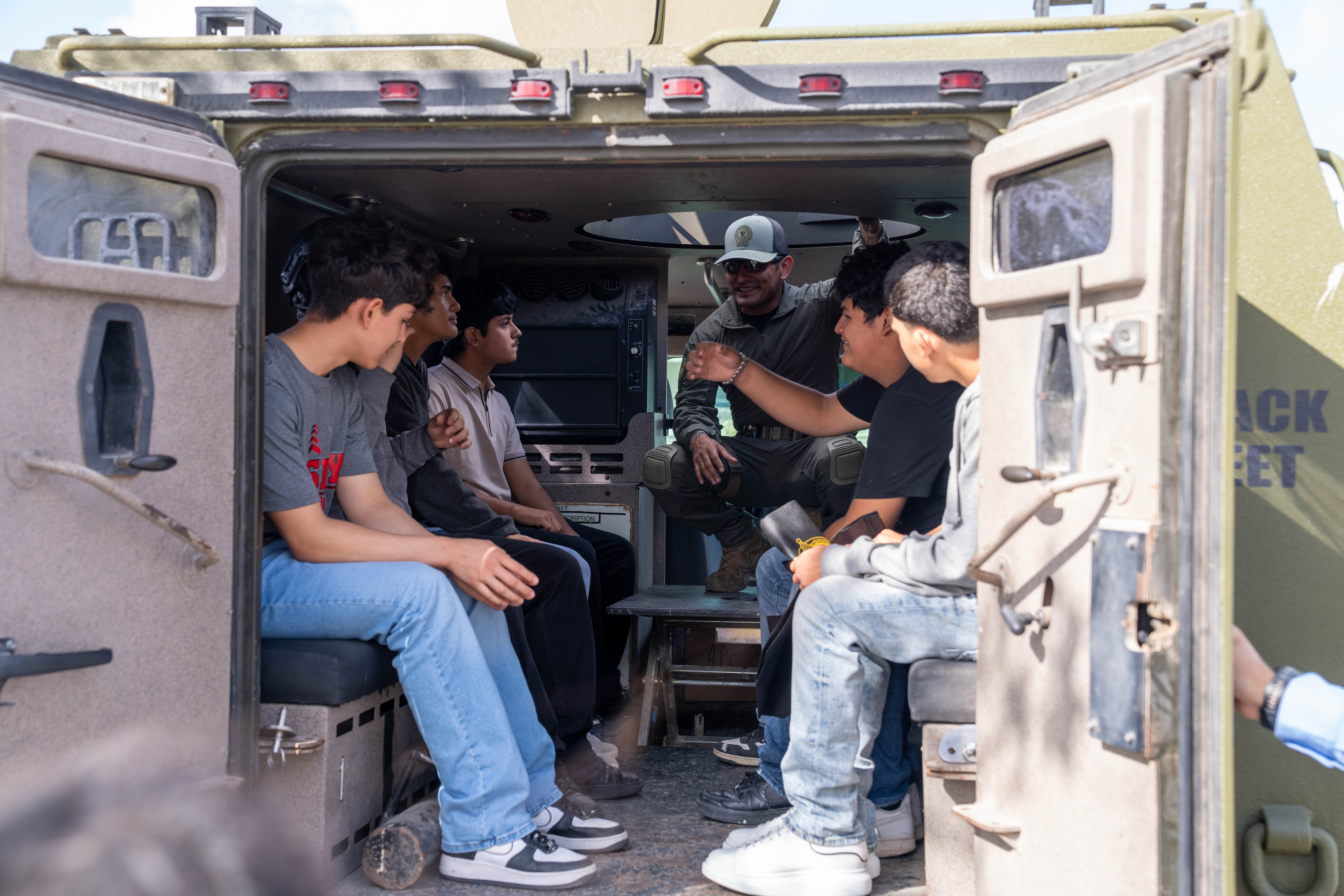 A man in a tactical uniform speaks to a group of young people seated inside an armored vehicle.