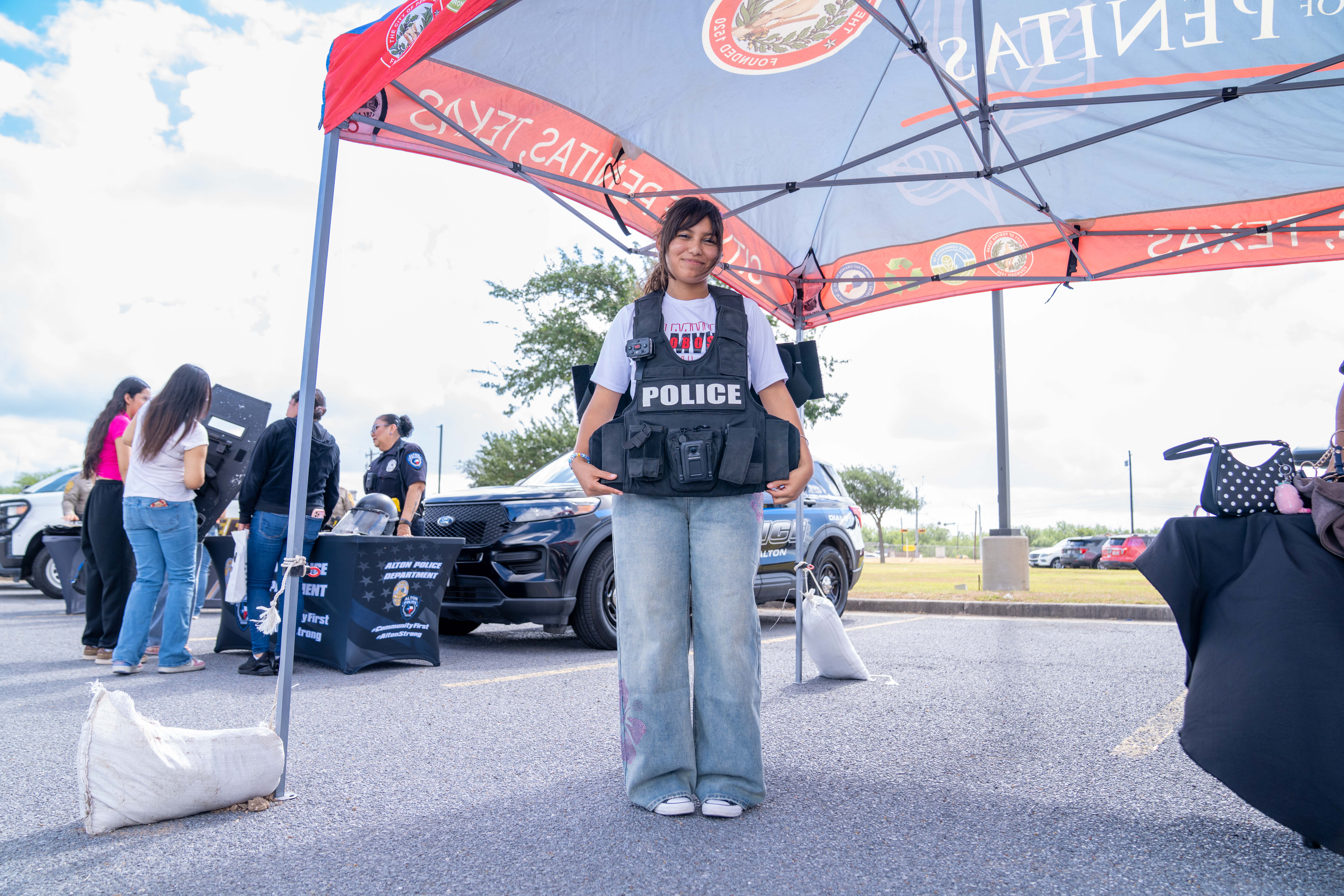 A young woman smiles while wearing a police vest at an outdoor event.