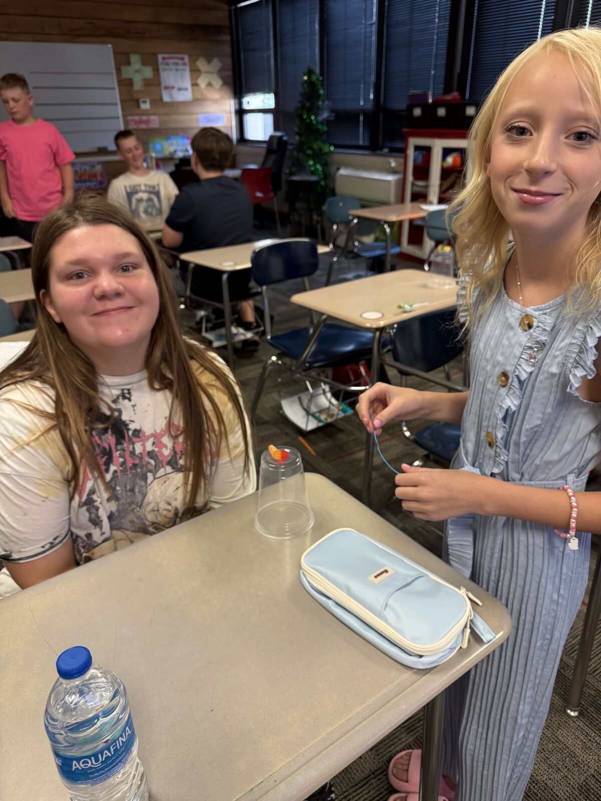Two young girls smile at the camera while sitting at desks in a classroom.