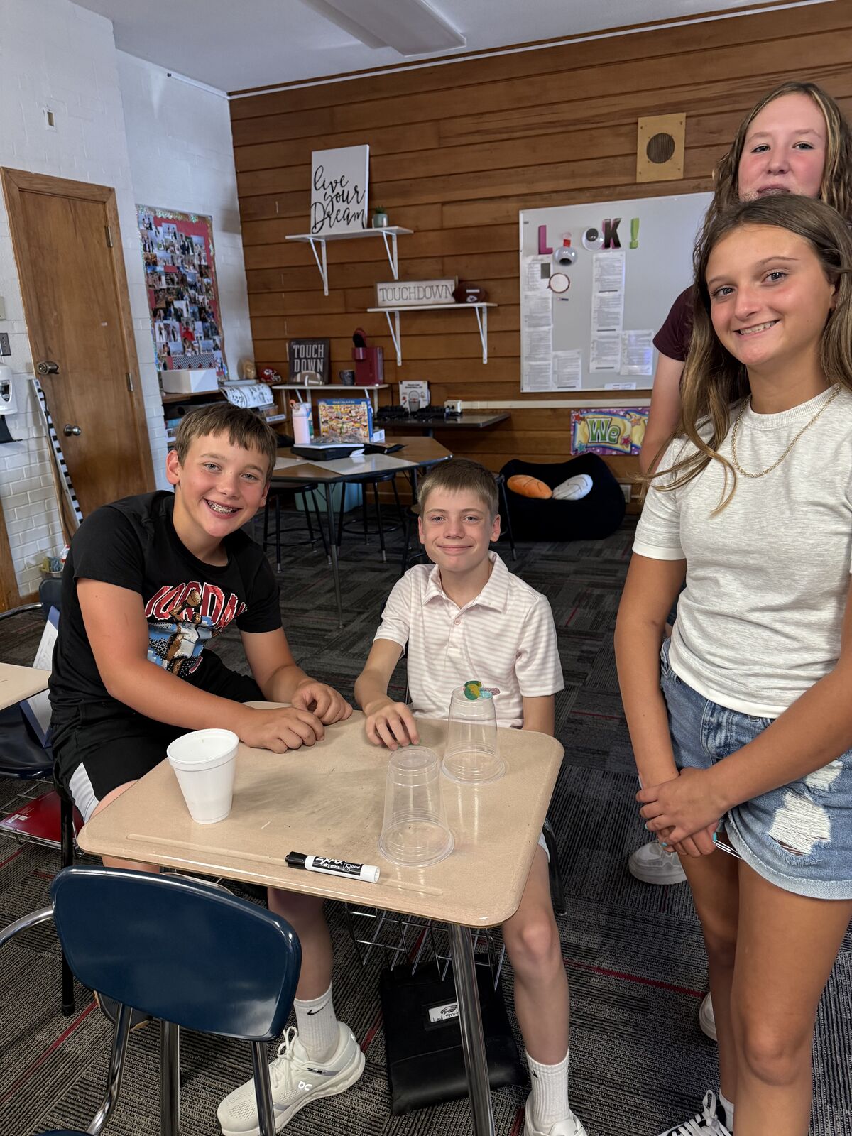 Three smiling students are gathered around a desk in a classroom, with two boys in the foreground and two girls behind them.
