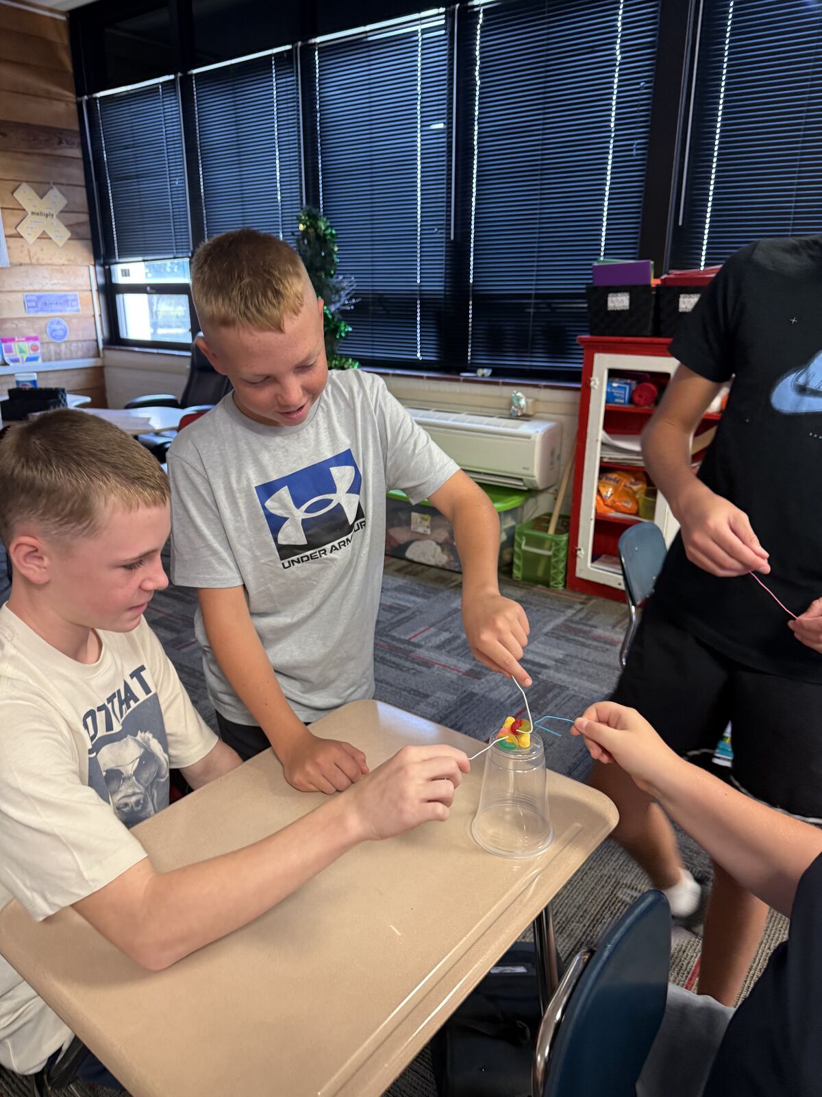Students work together on a science experiment involving a clear cup and colorful objects.