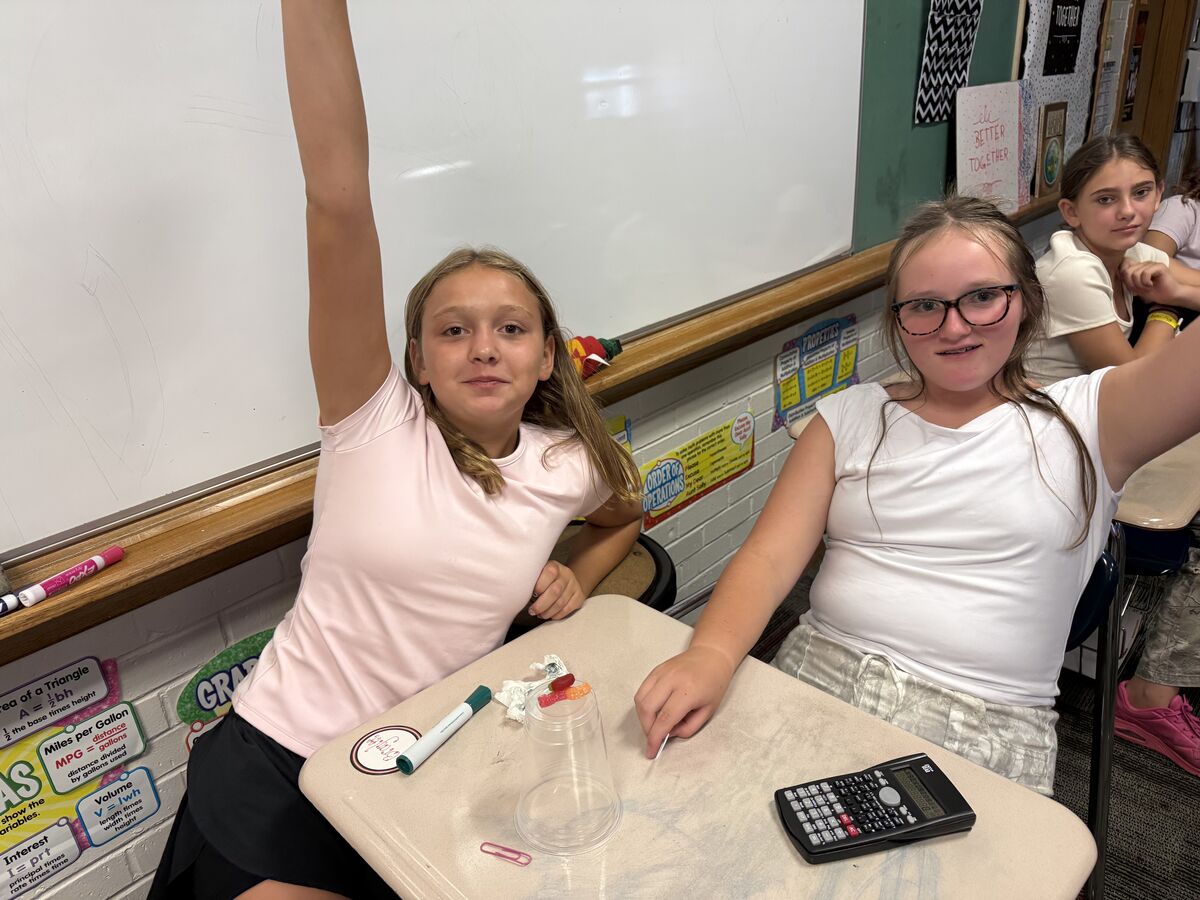 Two girls sit at desks in a classroom, with one raising her hand and the other smiling.