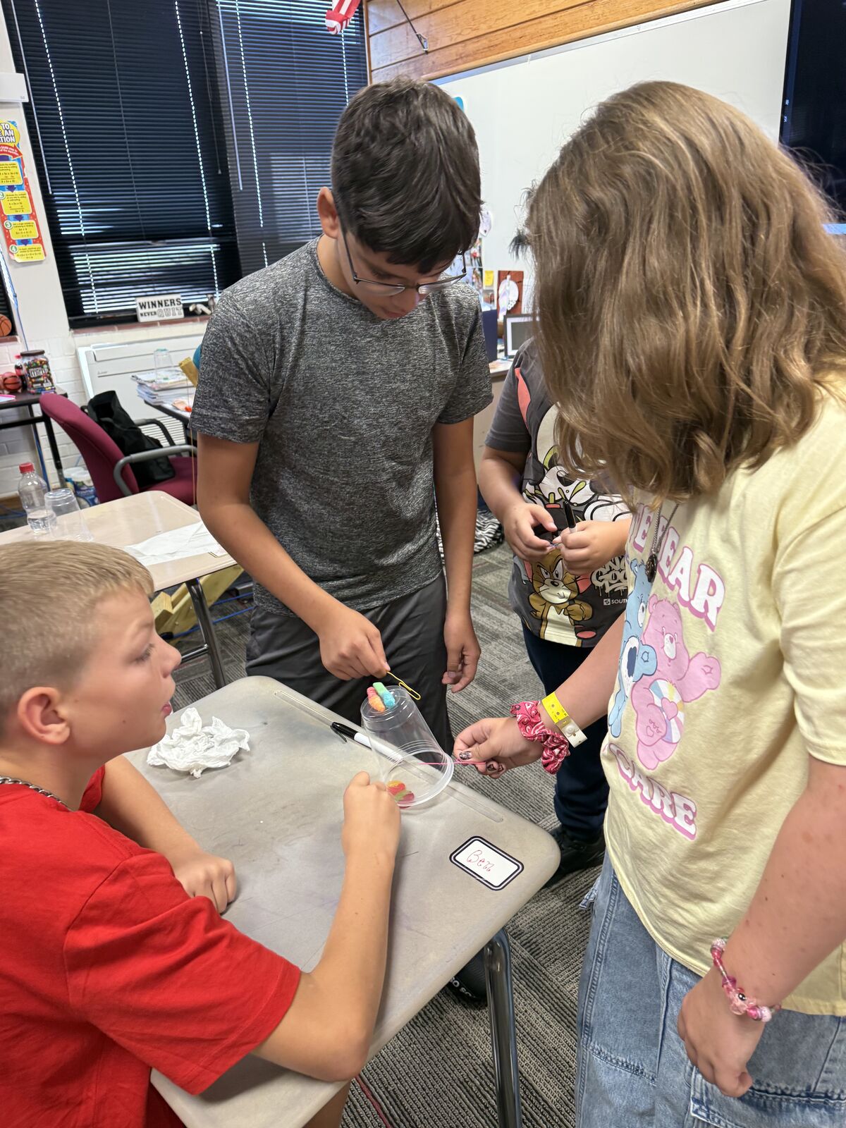 Children engage in a science experiment, using a clear cup and colorful gummy worms.