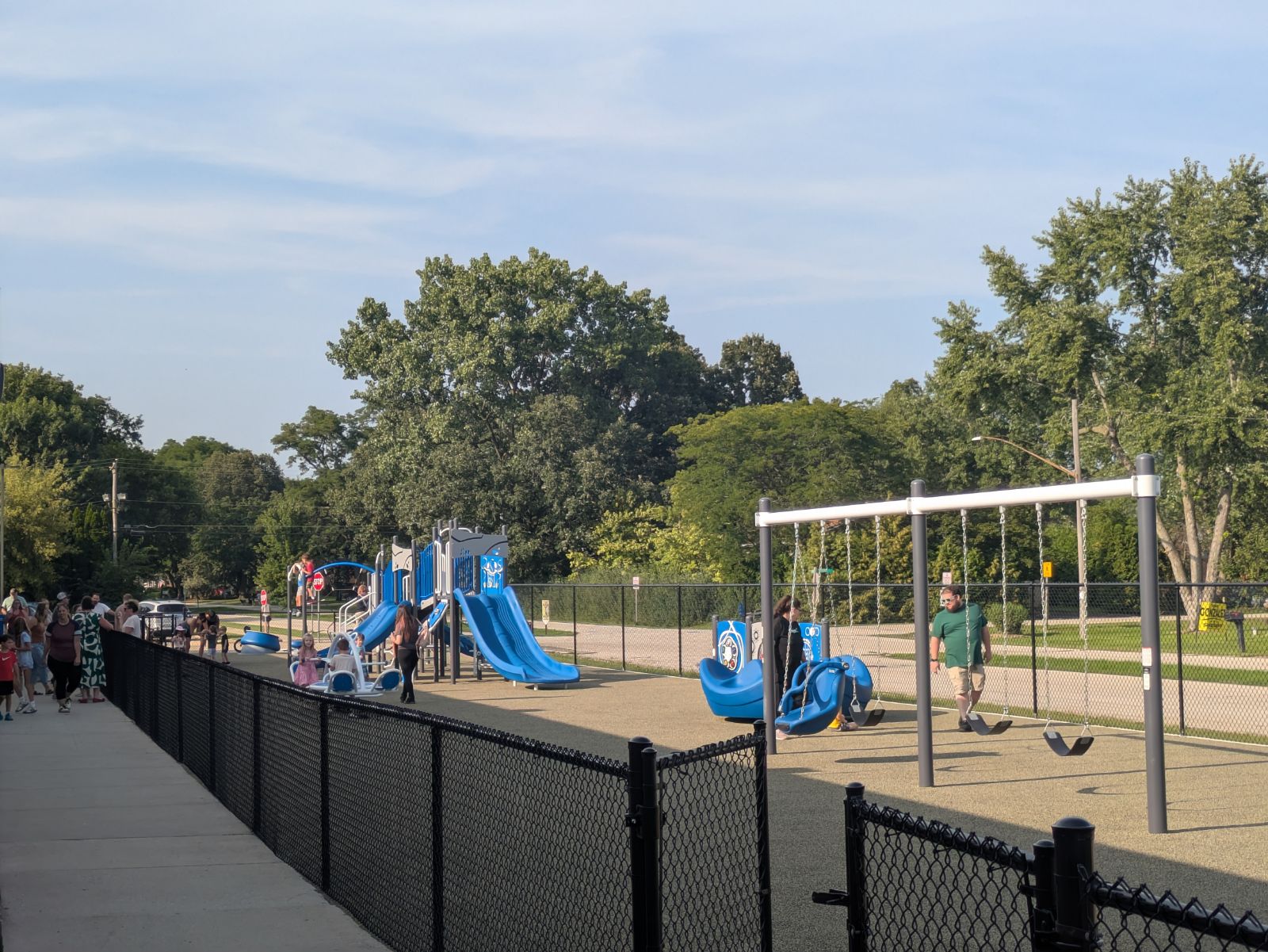 A bright blue playground with slides and swings is filled with children and adults on a sunny day.