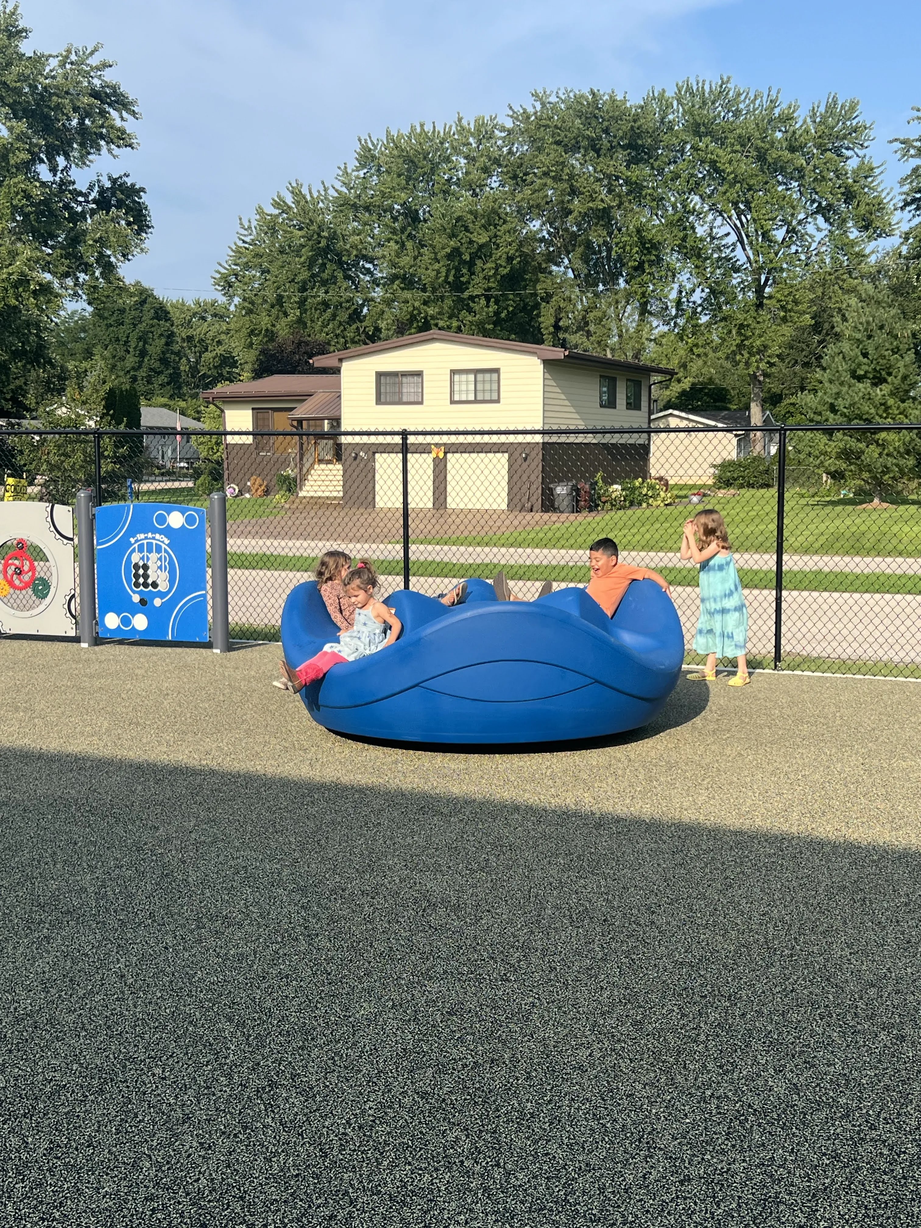 Children play on a blue spinning toy at a playground with a house and trees in the background.