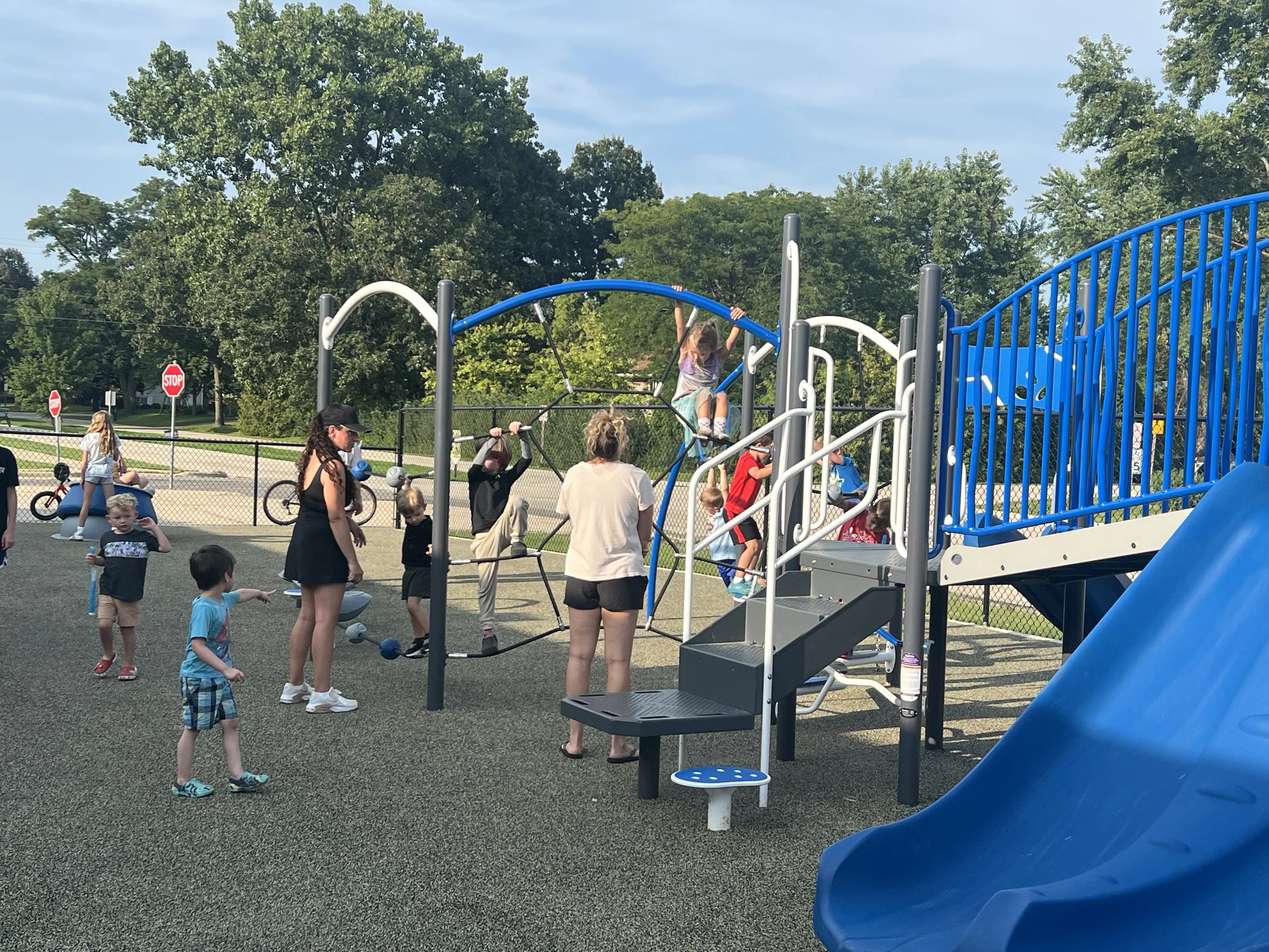 Children play on a colorful playground structure with slides and climbing nets, supervised by adults.