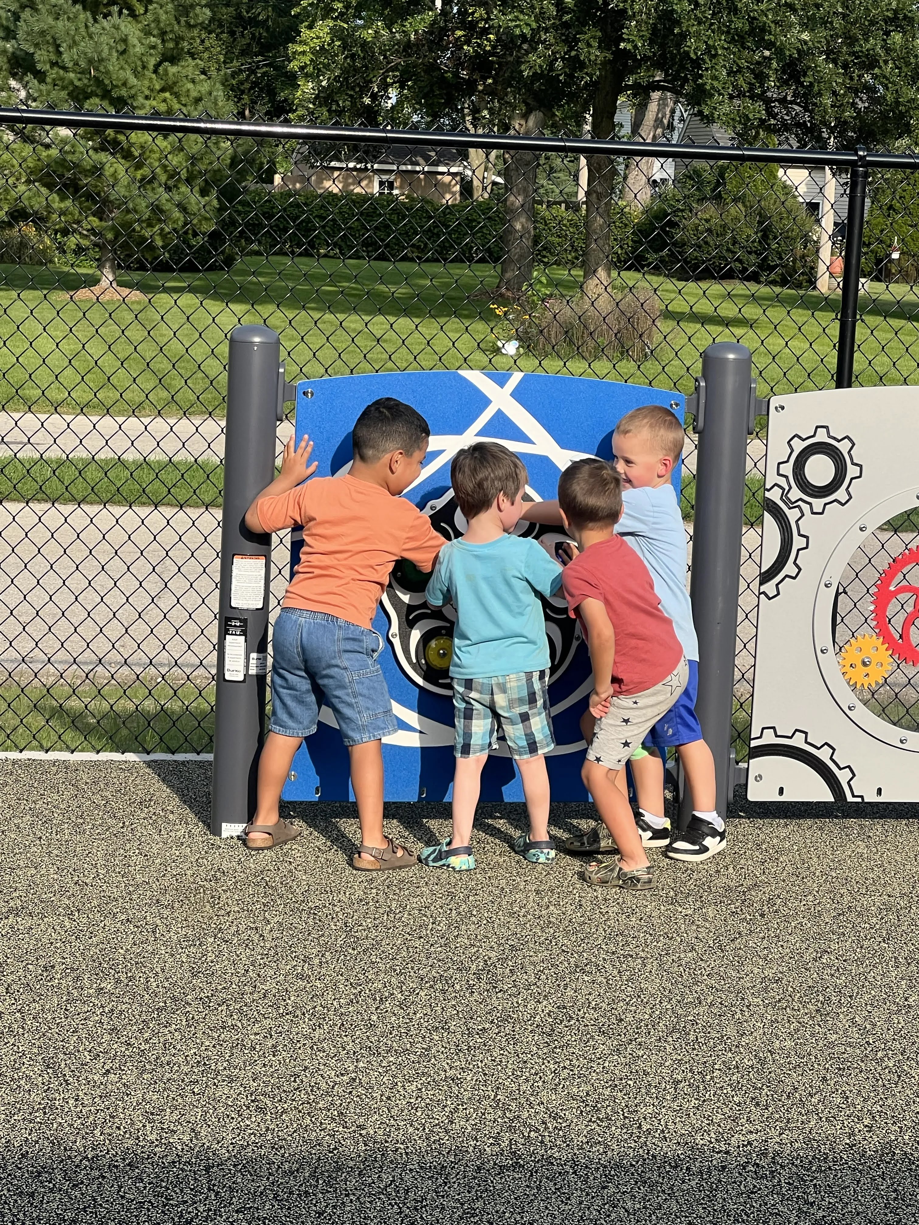 Four young boys interact with a colorful outdoor play panel featuring gears and interactive elements.