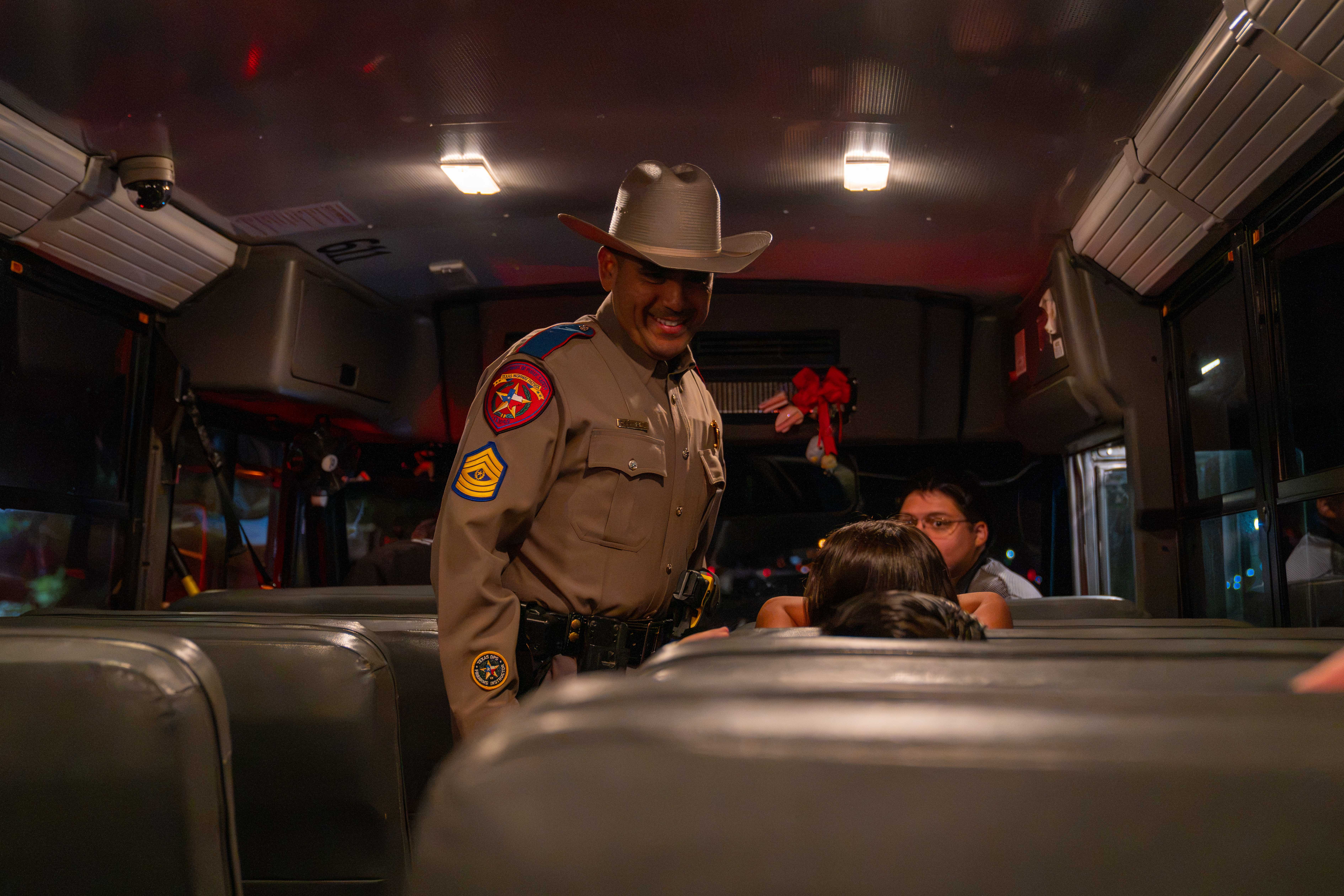 A smiling state trooper in uniform and a cowboy hat talks with passengers inside a bus.