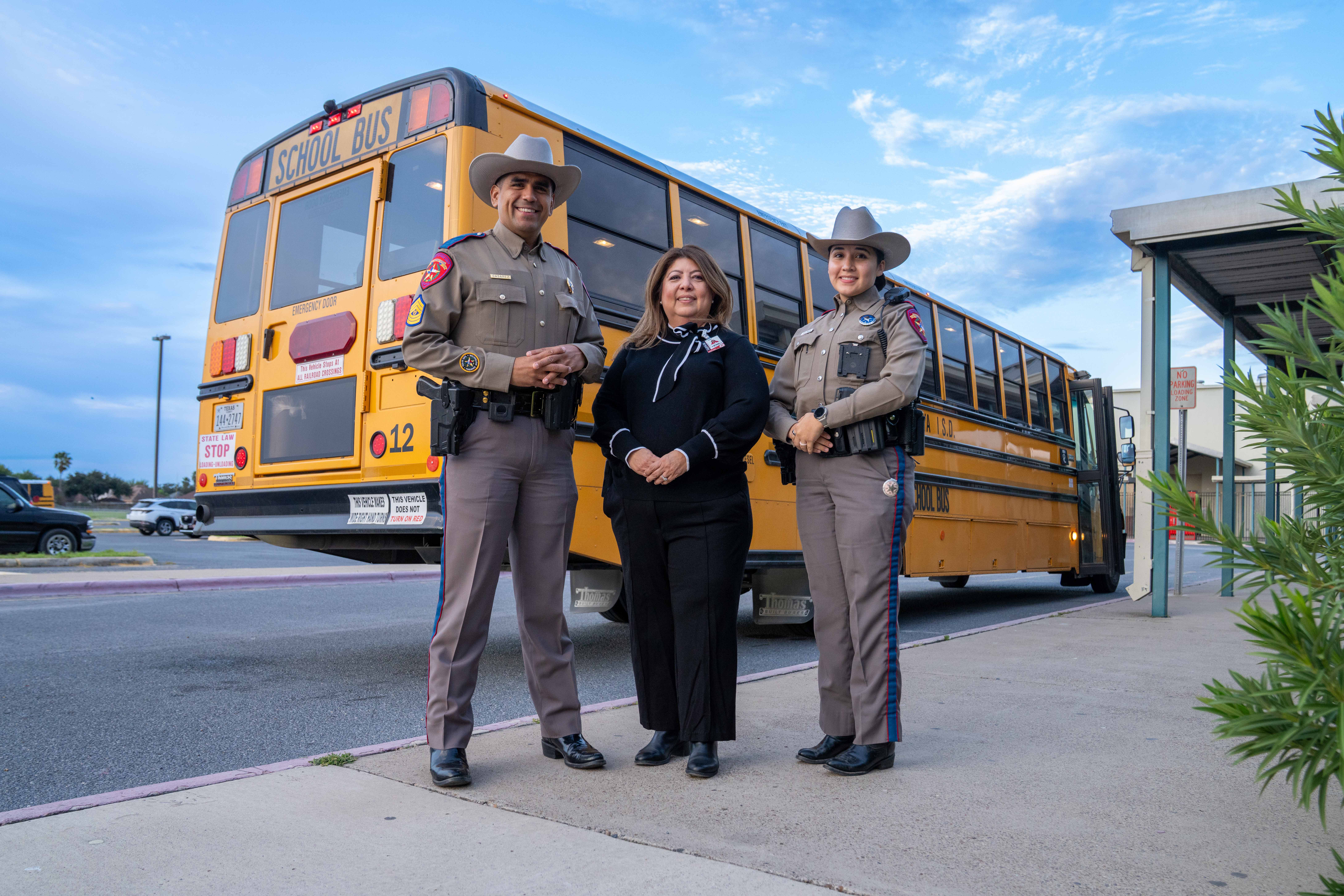 Two state troopers and a woman stand together on a sidewalk in front of a yellow school bus outdoors.