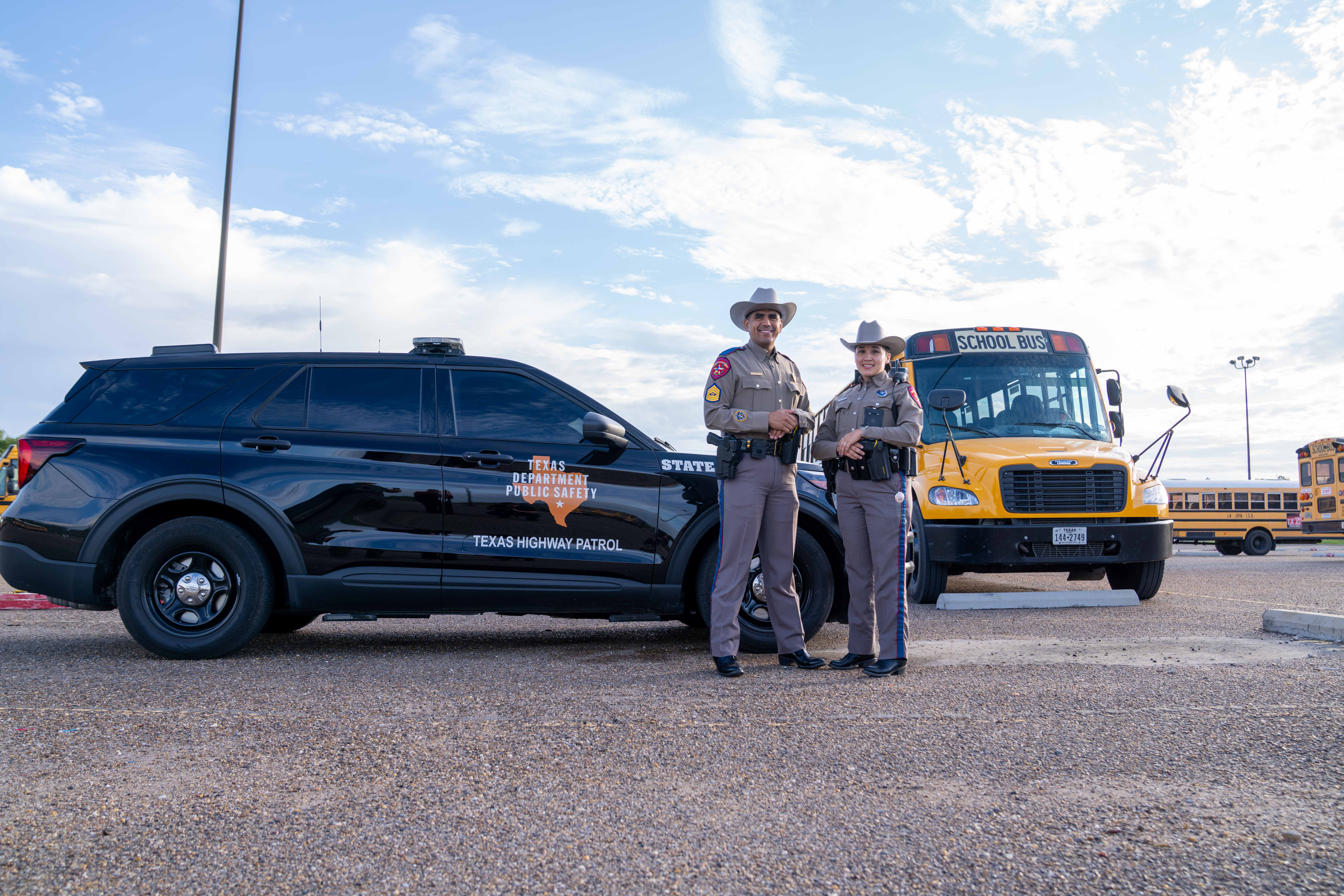 Two Texas Highway Patrol officers stand proudly between a black patrol SUV and a yellow school bus outdoors.