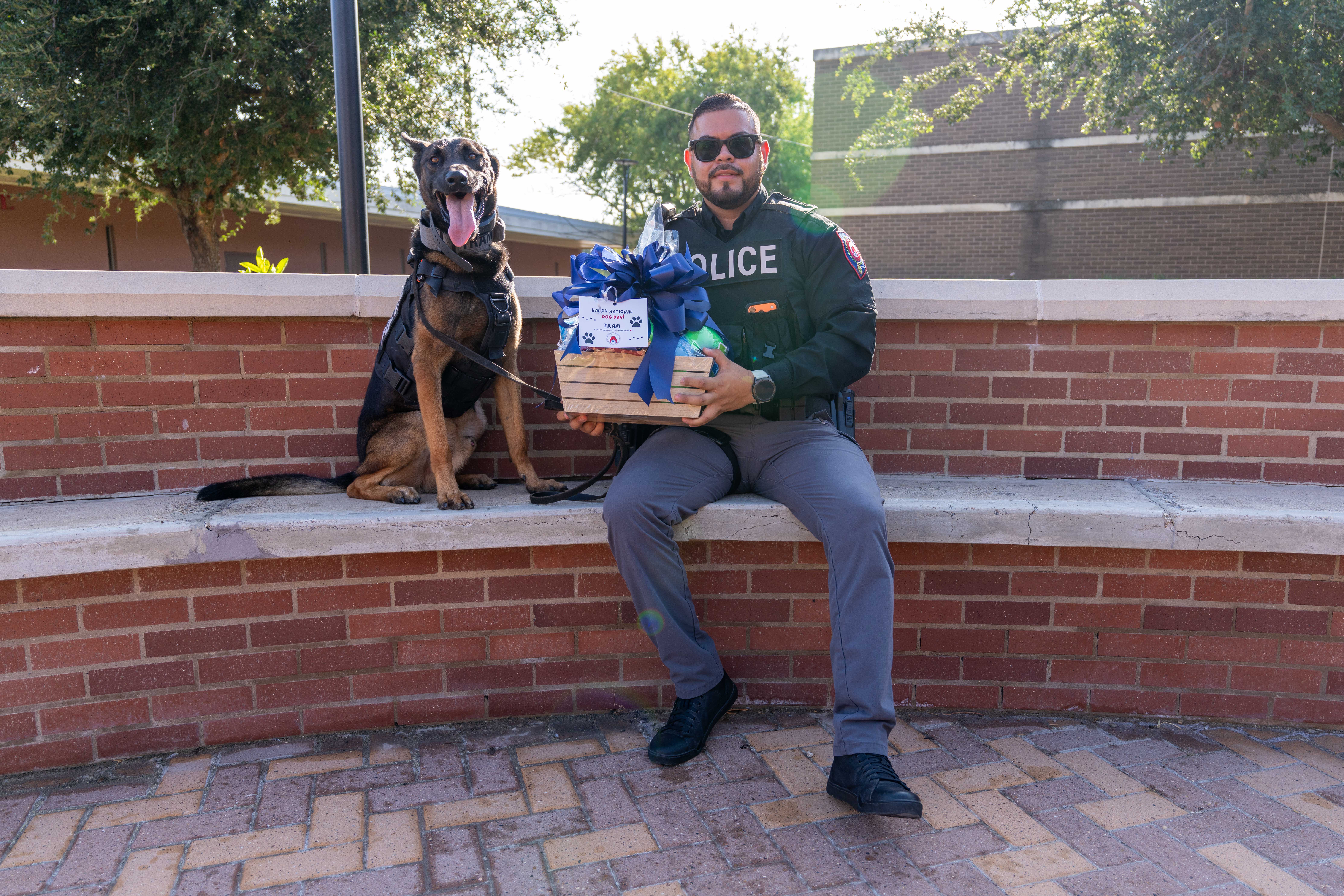 A smiling police officer in uniform sits on a brick wall holding a gift basket, accompanied by his K9 dog sitting beside him.