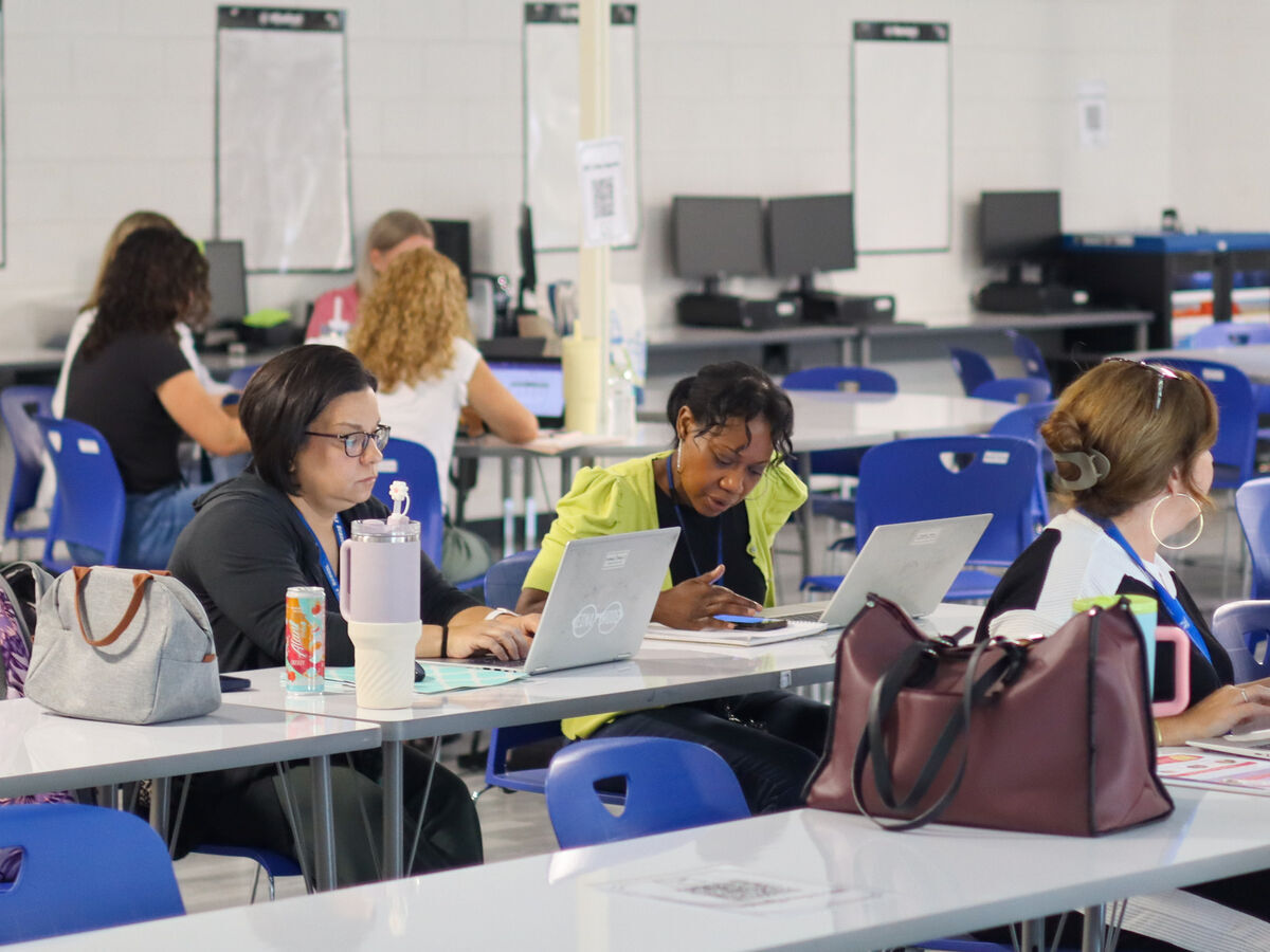 Several people are seated at tables in a classroom or conference setting, each working on a laptop.