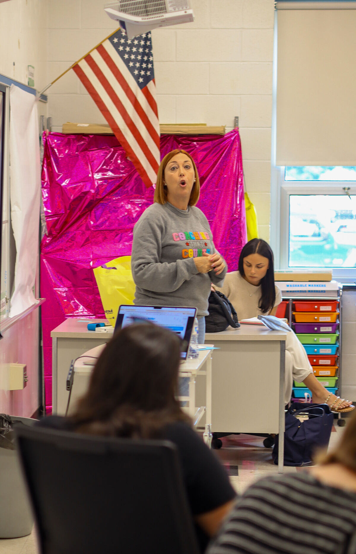 A woman in a grey sweatshirt with colorful text stands and speaks in a classroom.