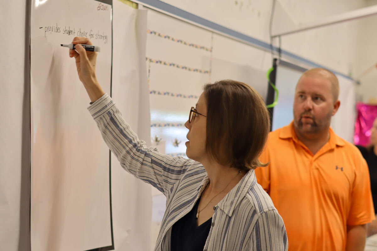 A woman with glasses writes on a whiteboard with a marker, while a man in an orange shirt looks on.
