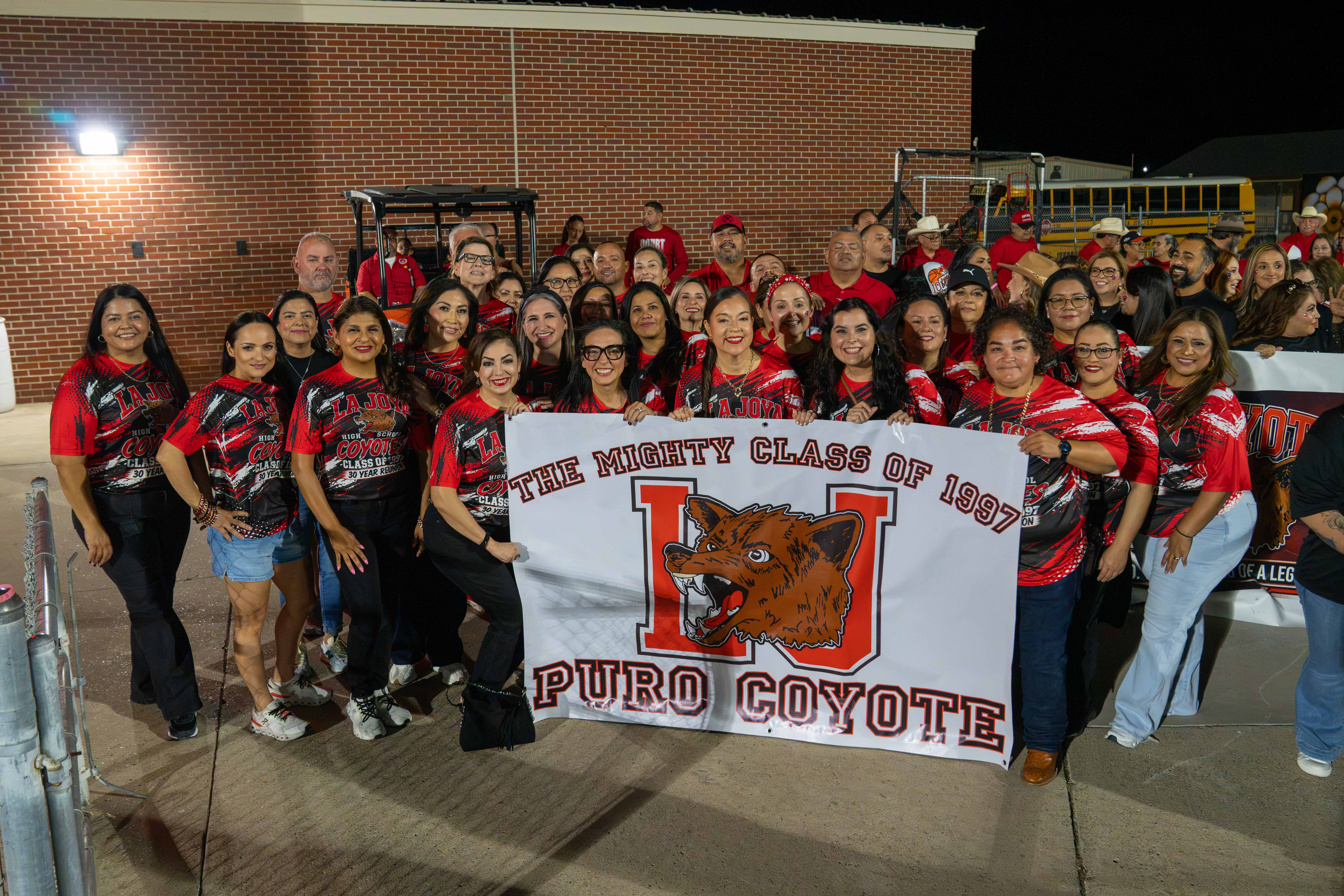 A large group of smiling adults gathers outdoors at night, holding a banner that reads "The Mighty Class of 1997 Puro Coyote".