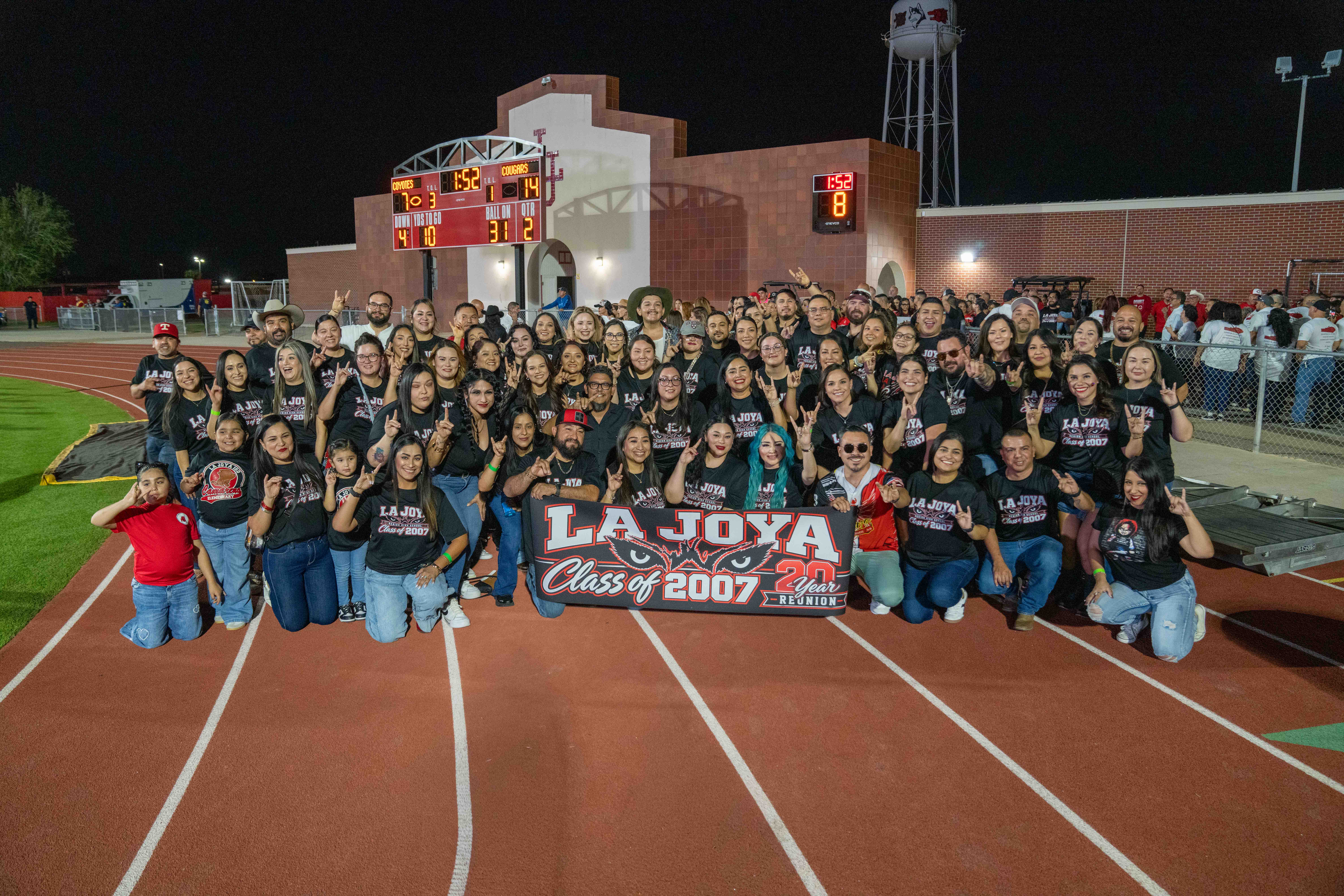 A large group of adults gathers on a running track at night for a high school reunion, holding a banner that reads "LA JOYA Class of 2007 20 Year REUNION."