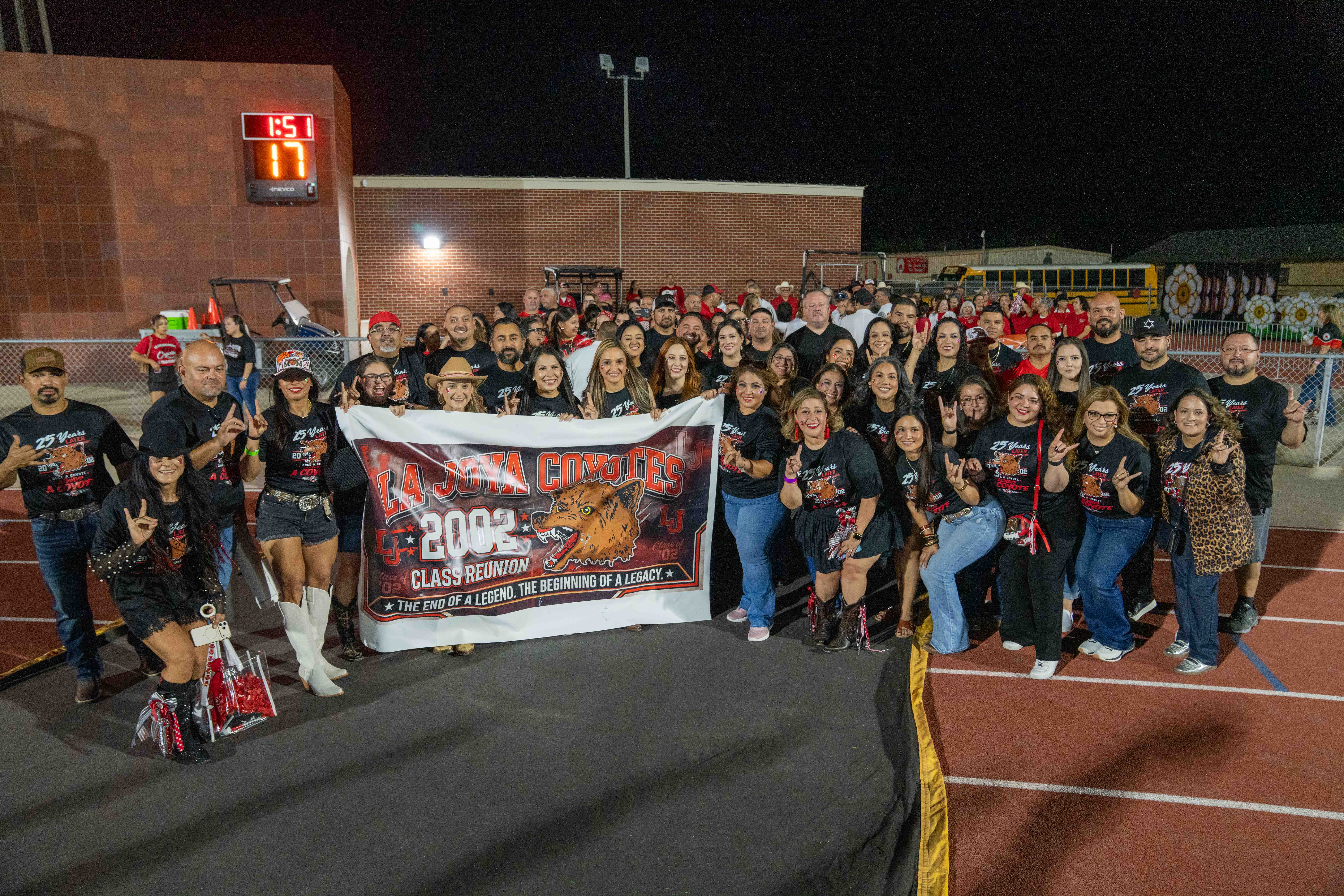 A group of people gathers on a running track at night, holding a large banner that reads "La Joya Coyotes Class Reunion 2002."