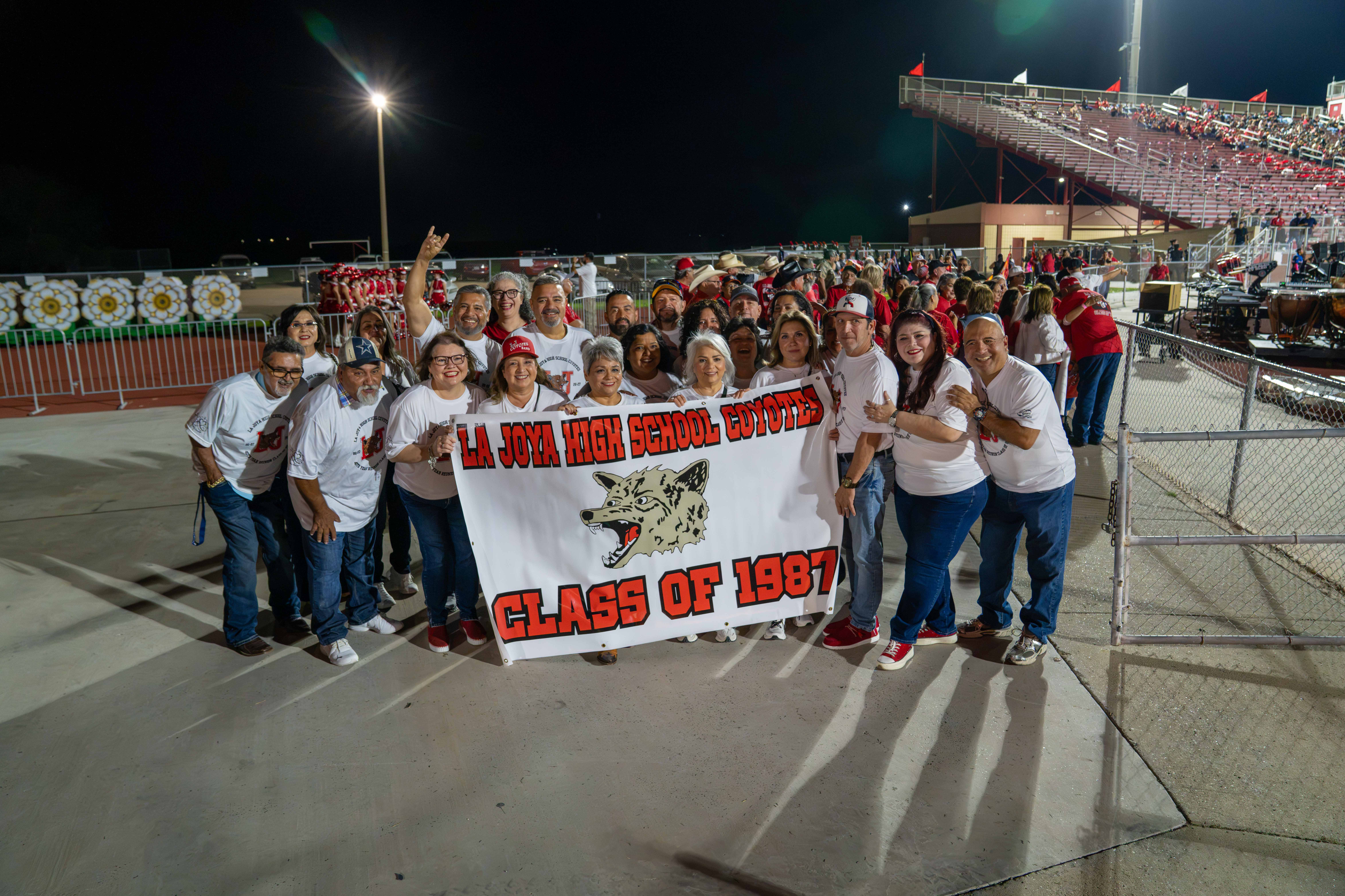 A group of alumni hold a banner reading "LA JOYA HIGH SCHOOL COYOTES CLASS OF 1987" at a night event in a stadium.
