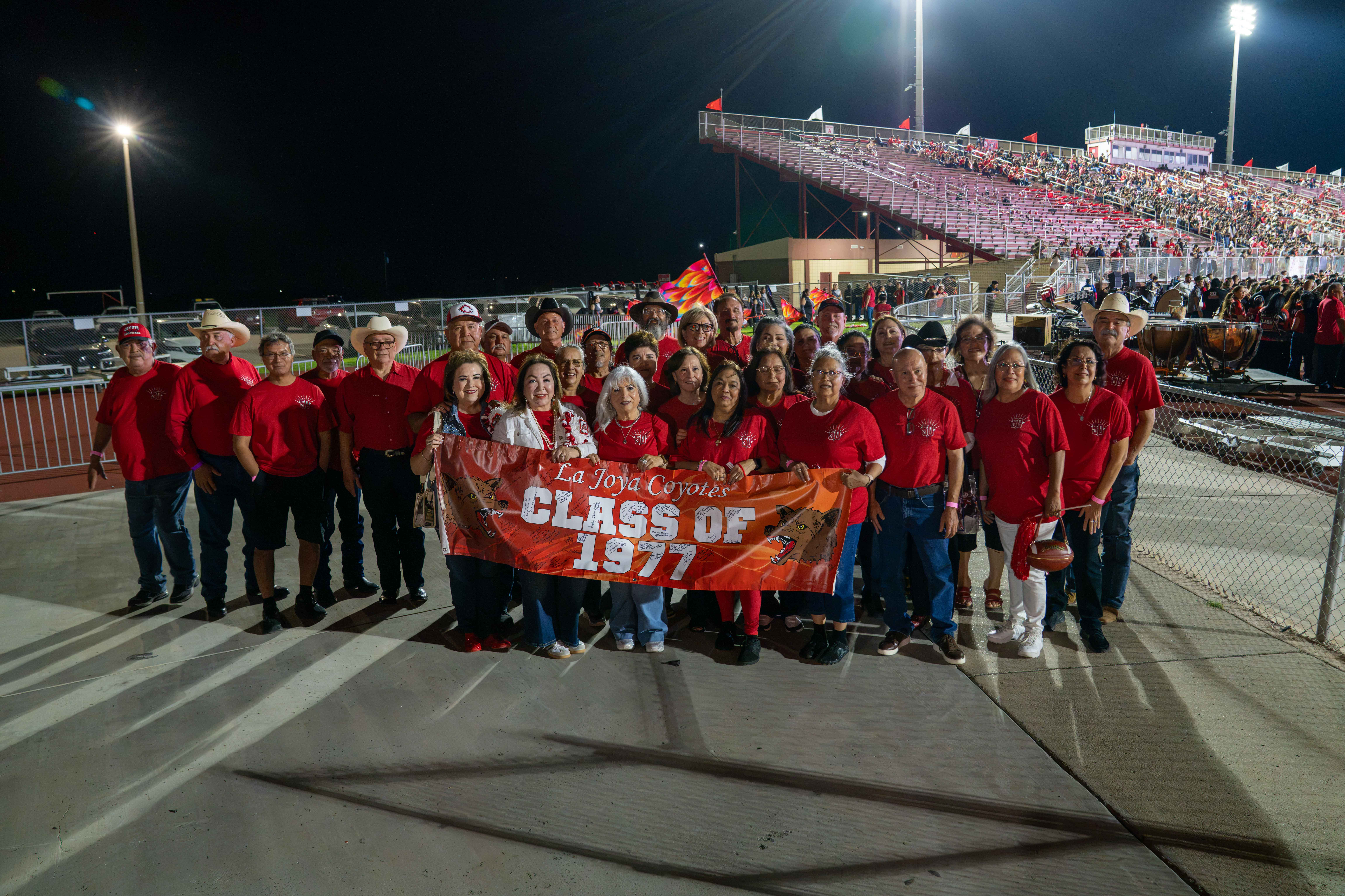 A group of alumni from the La Joya Coyotes Class of 1977 gathers at night outside a stadium, holding a banner and wearing matching red shirts.