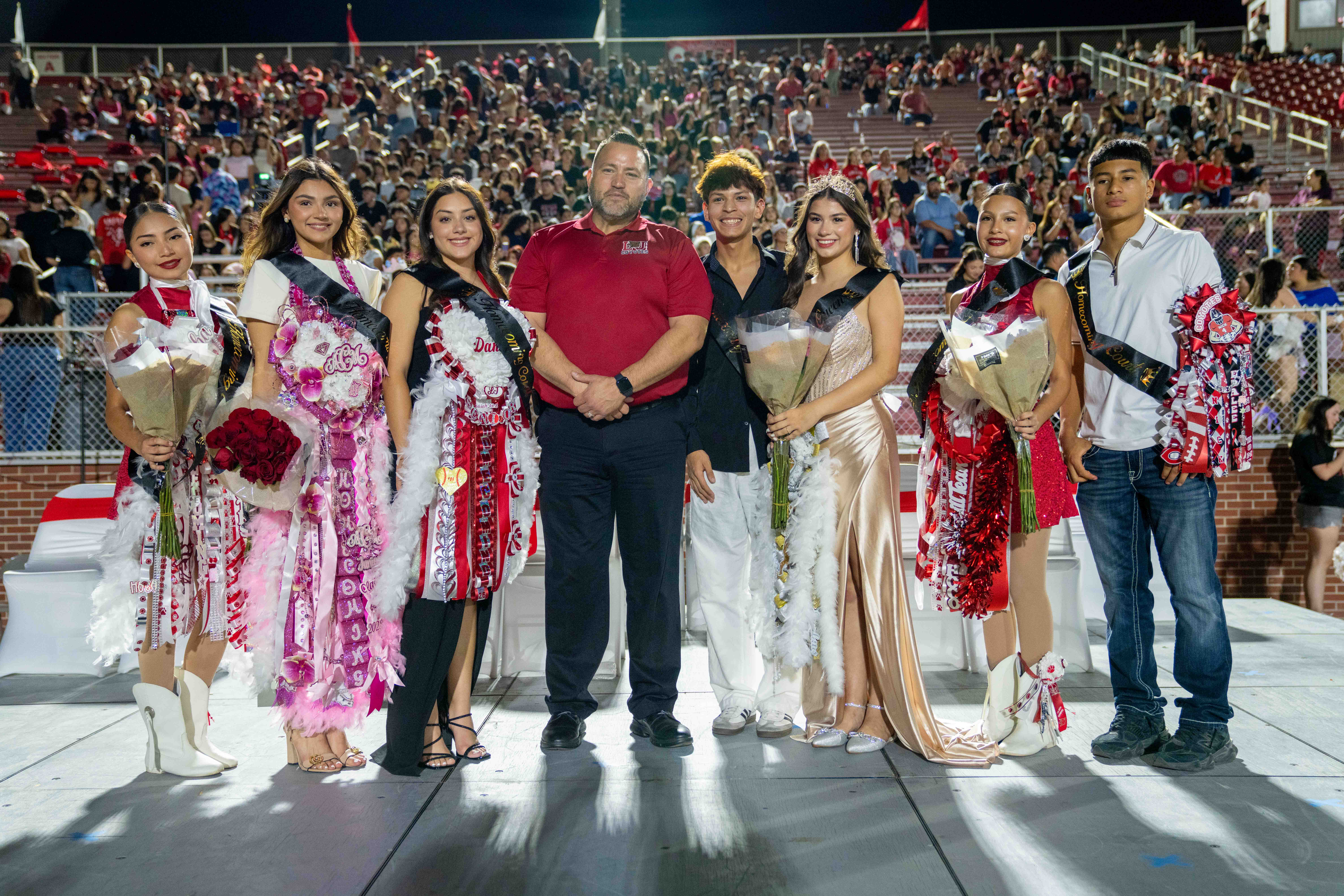 A group of seven students and an adult stand proudly on a stage in front of a packed stadium bleacher at night, posing for a homecoming photo.