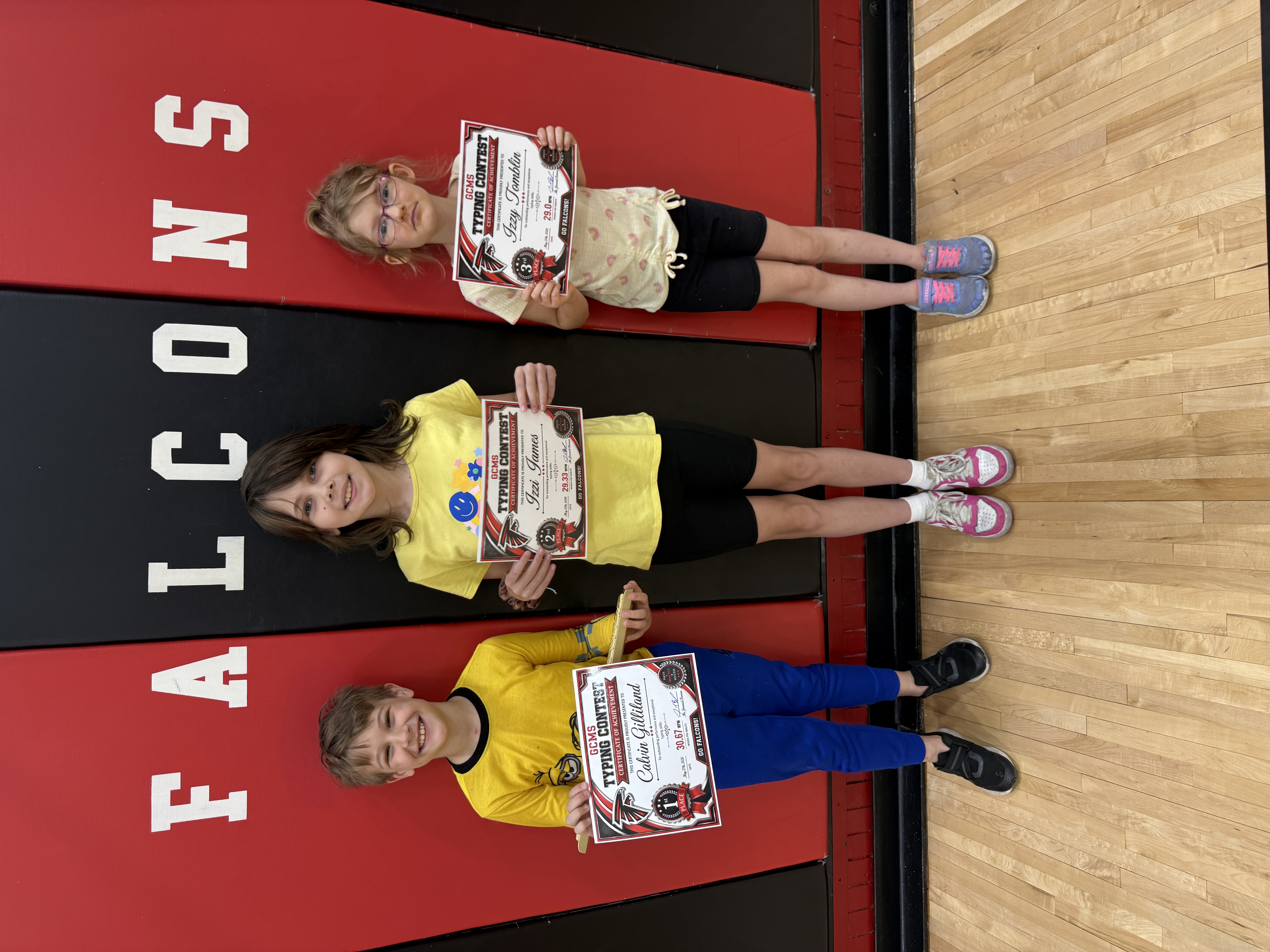 Three children proudly display their typing contest certificates on a red and black mat with "FALCONS" printed on it.