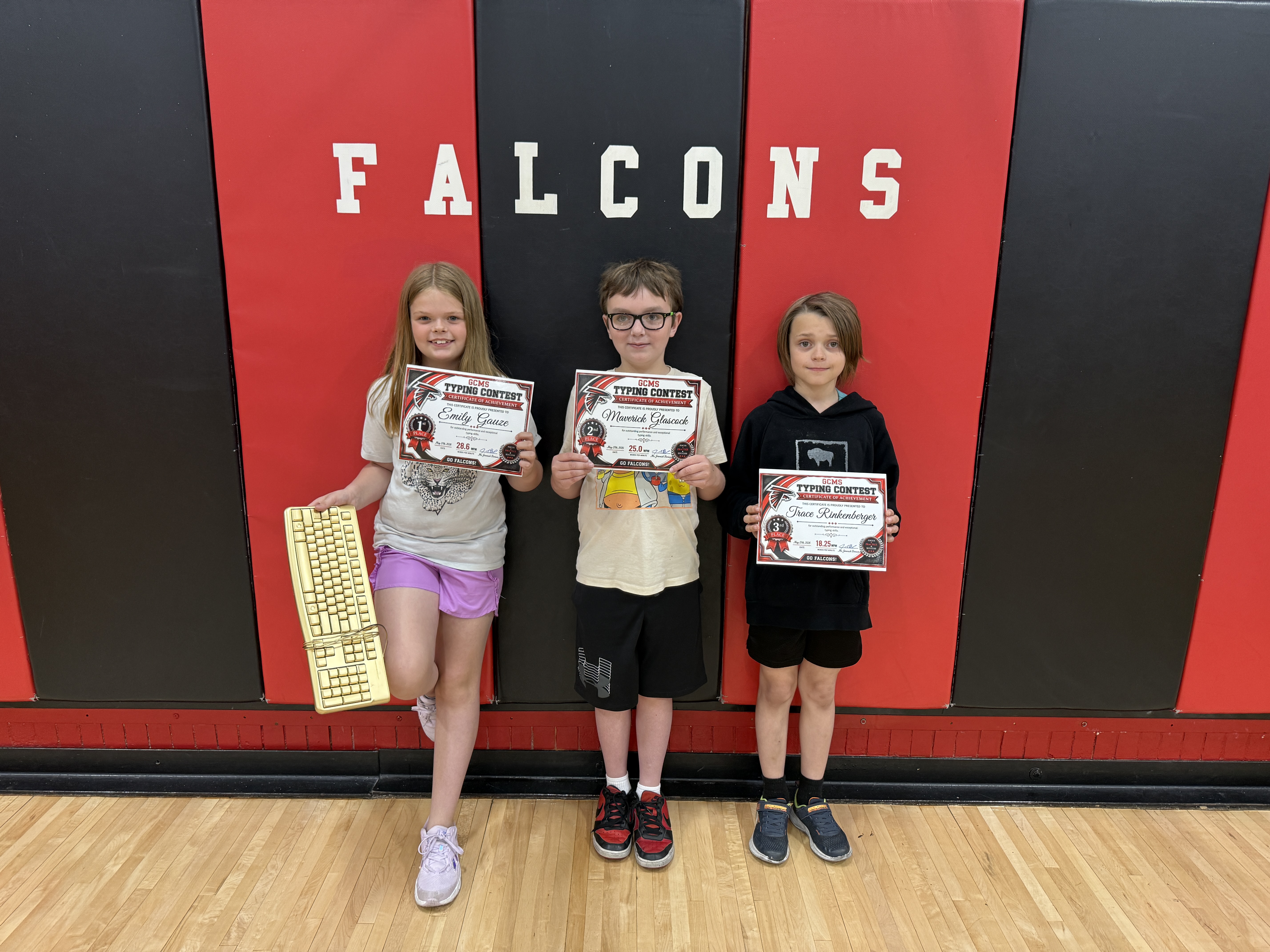 Three students stand in front of a 'FALCONS' banner, holding certificates for a typing contest.