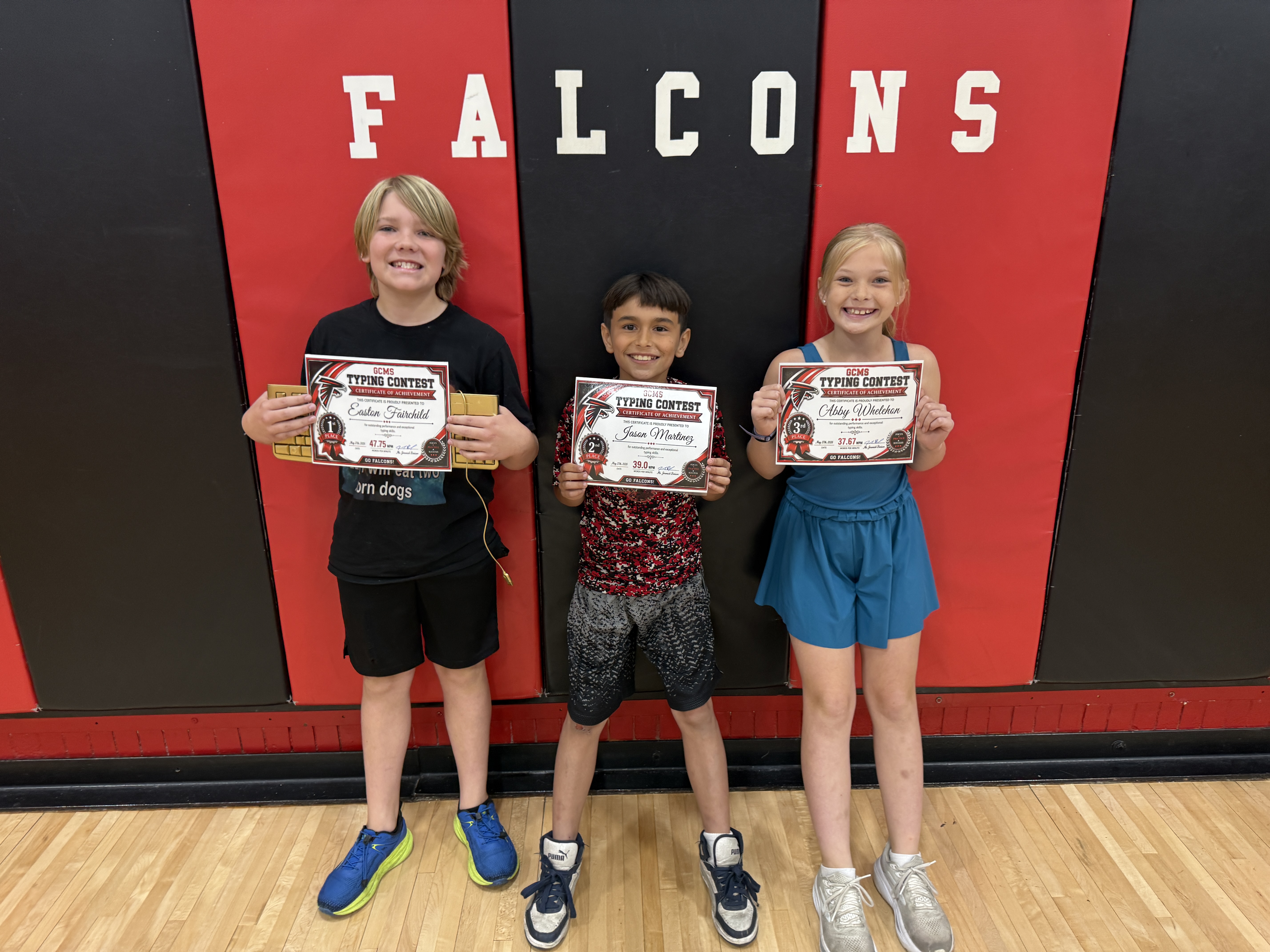 Three smiling children hold certificates for a typing contest in front of a wall with 'FALCONS' written on it.