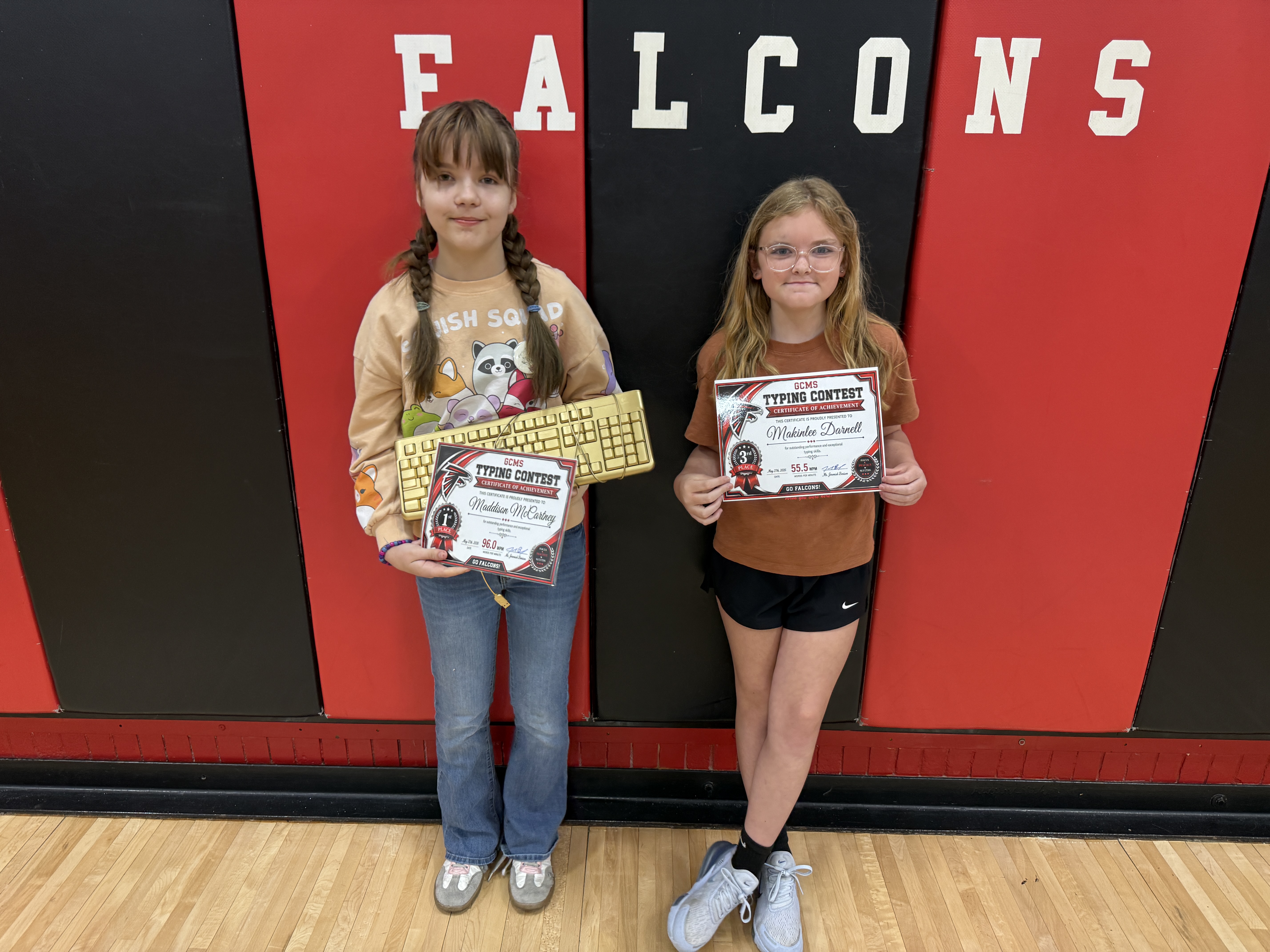 Two girls stand in front of a wall with "FALCONS" written on it, holding certificates and keyboards from a typing contest.