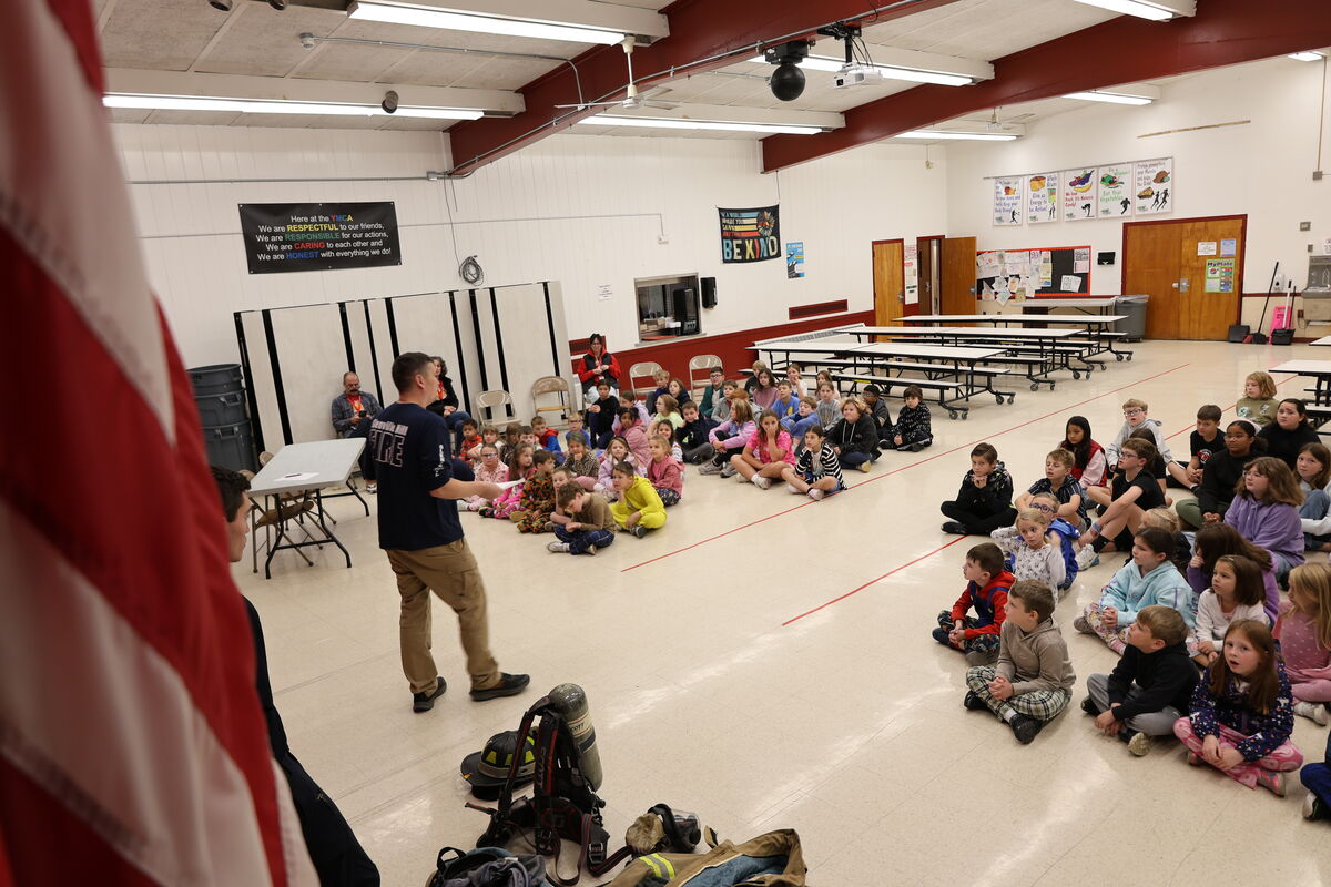 A speaker in a dark blue shirt presents to a group of children sitting on a school cafeteria floor.