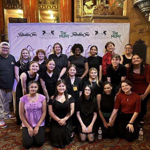 A diverse group of young people and adults pose for a photo in front of a banner with logos for Fabulous Fox, Fox Performing Arts Charitable Foundation, and The Muny.