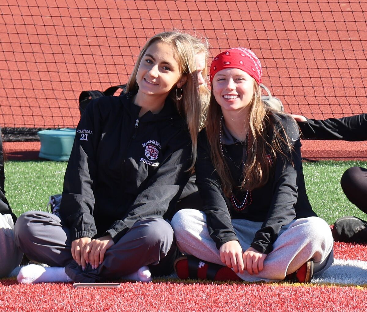 Two young women sit cross-legged on a sports field, smiling for a sunny outdoor photo.