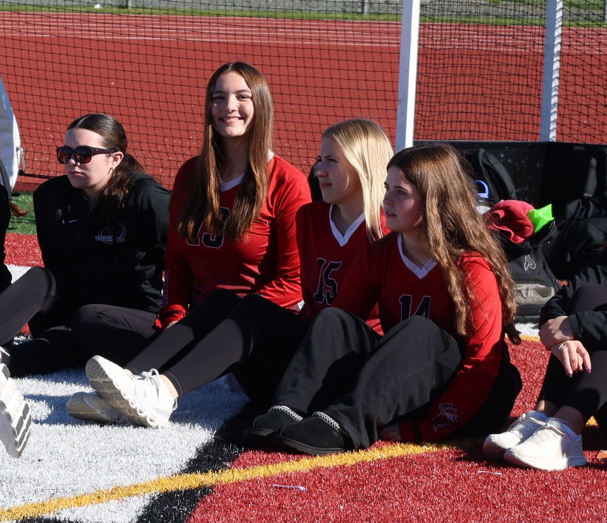 Four young women in athletic wear sit together on a turf field, watching a game.