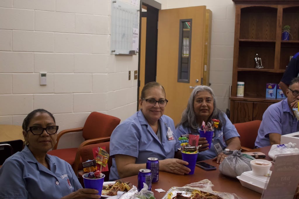 Three women in blue shirts sit at a table holding blue cups filled with candy bouquets.