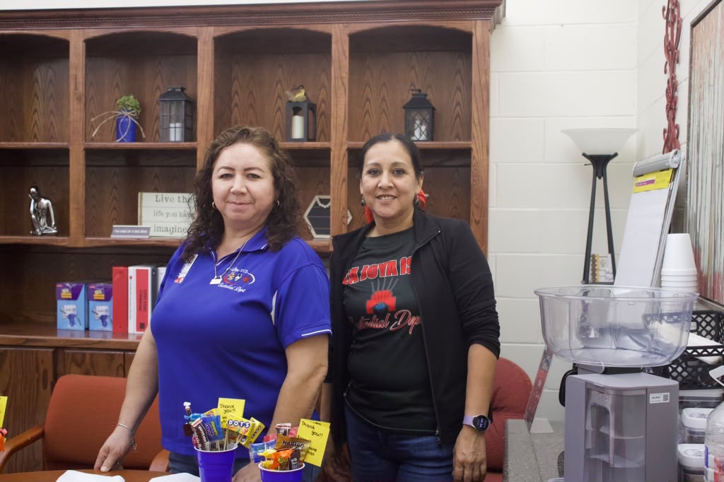 Two women stand together smiling in an office with large wooden shelves and a coffee maker.