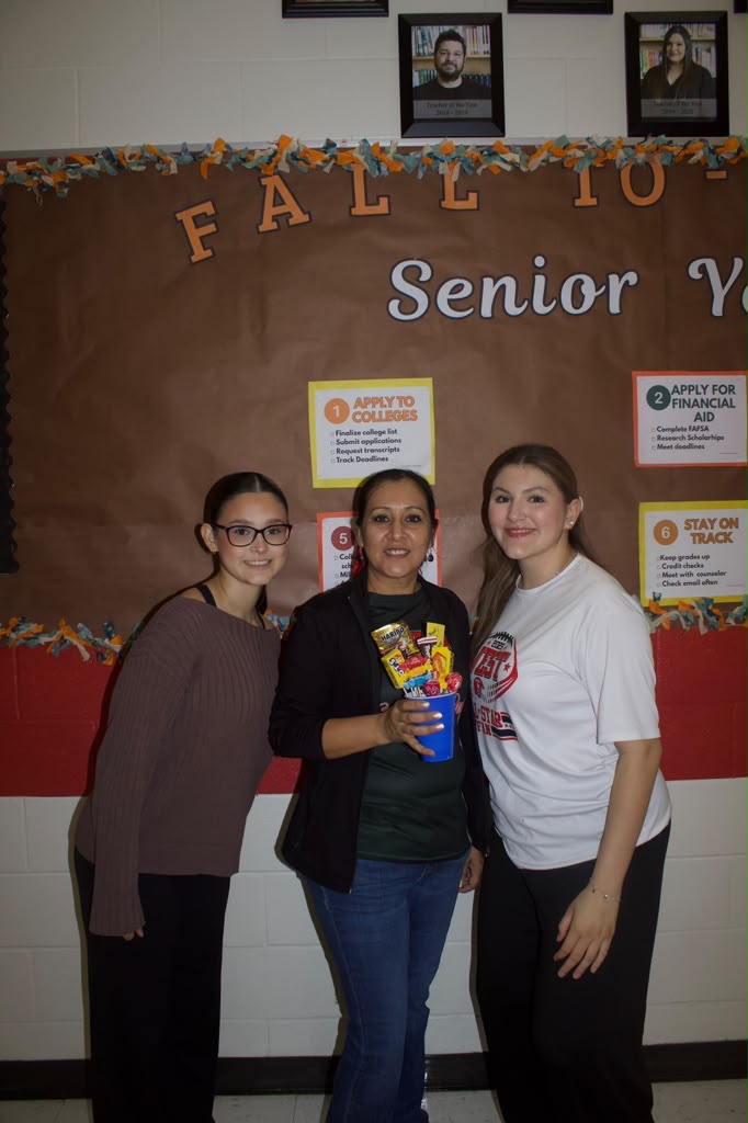 Three women stand smiling in front of a bulletin board with college preparation information.