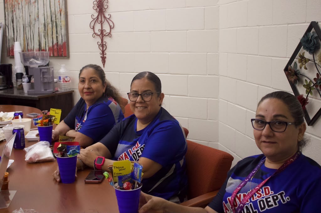 Three women in blue shirts sit at a long wooden table indoors, smiling at the camera.