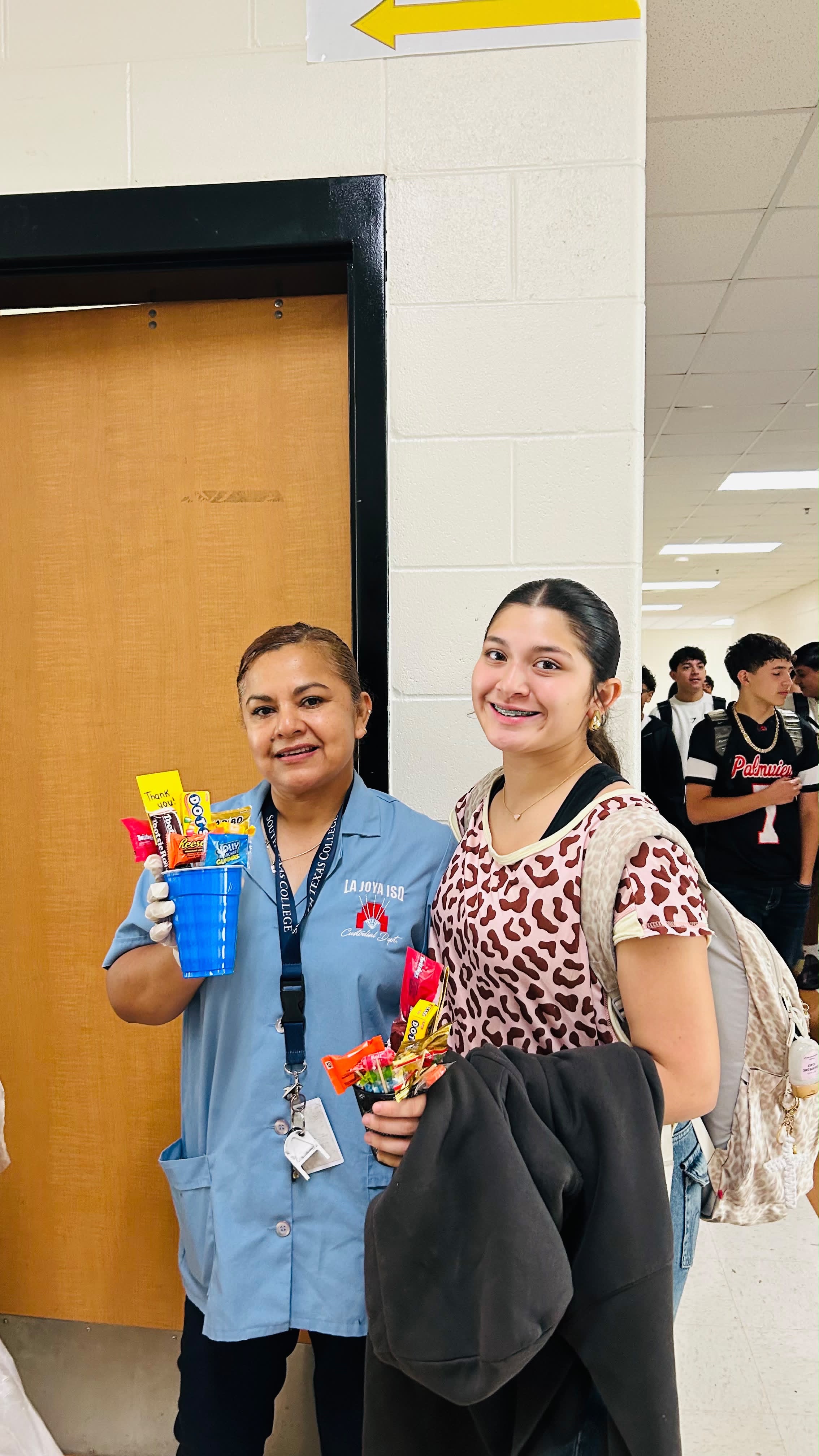 A smiling school custodian in blue uniform holds a blue cup filled with candy, standing beside a student in a leopard print shirt holding treats.