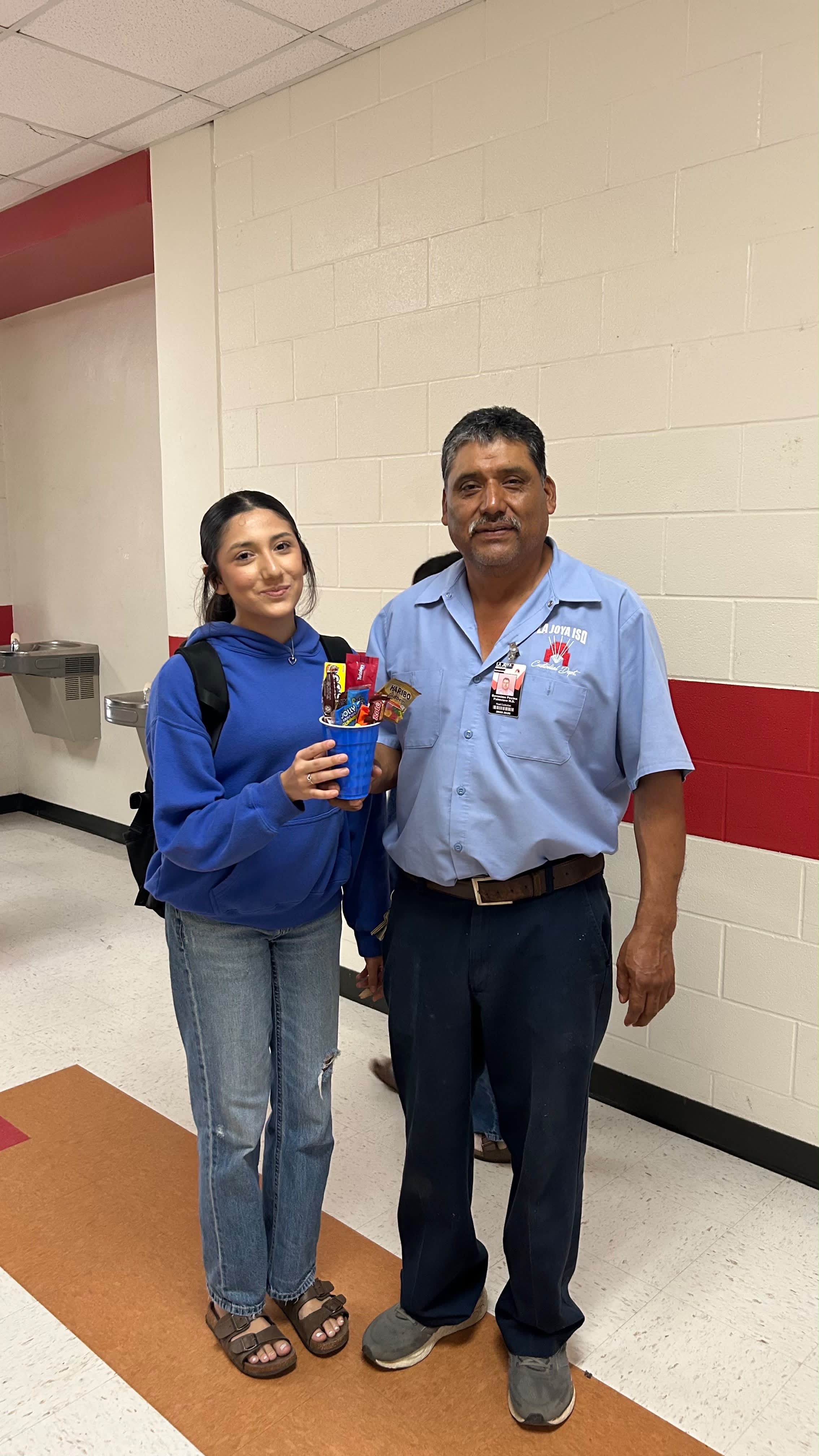 A student and a school staff member stand side by side in a hallway, holding a blue cup filled with candy.