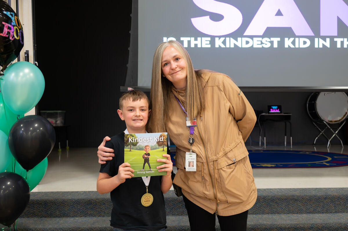A young boy proudly holds a book titled "Kindest Kid in the World" and a medal, standing next to a smiling woman.