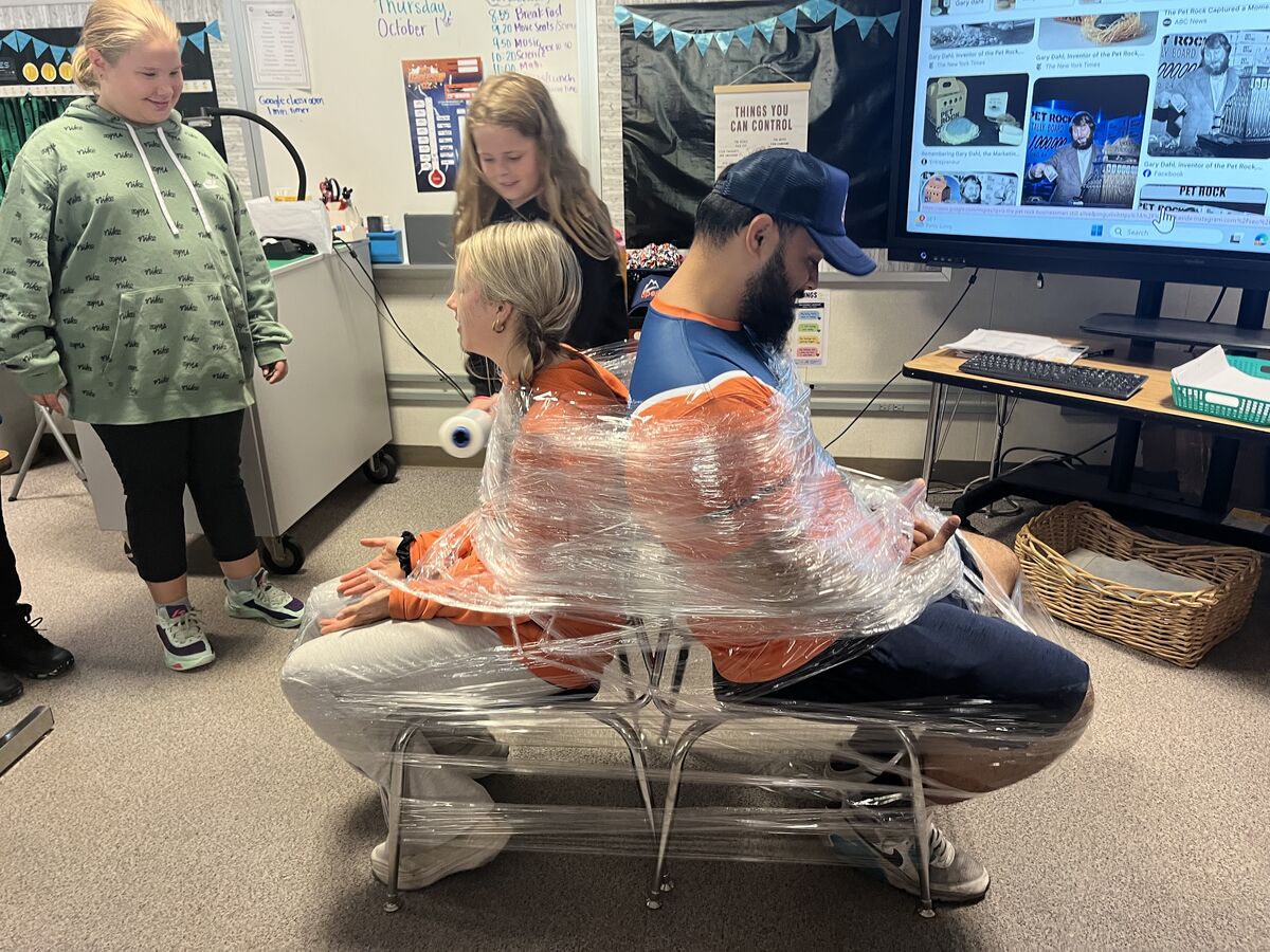 A woman and a man are wrapped together back-to-back in clear plastic wrap while sitting on chairs in a classroom.