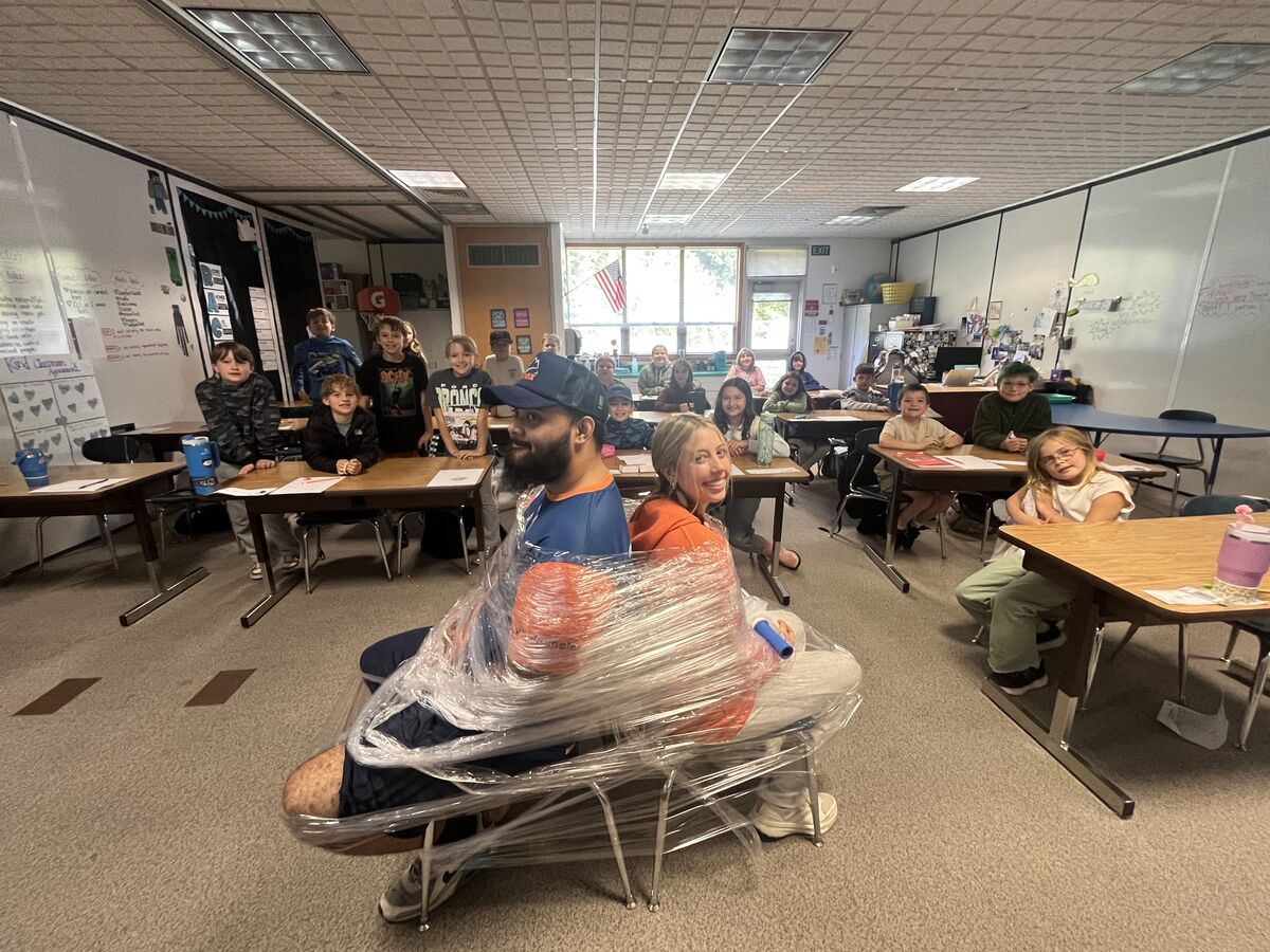 A man and woman are taped back-to-back in chairs by plastic wrap, in the middle of a classroom full of students watching.