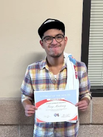 A young man in a plaid shirt and baseball cap proudly holds a "Student of the Month" certificate.