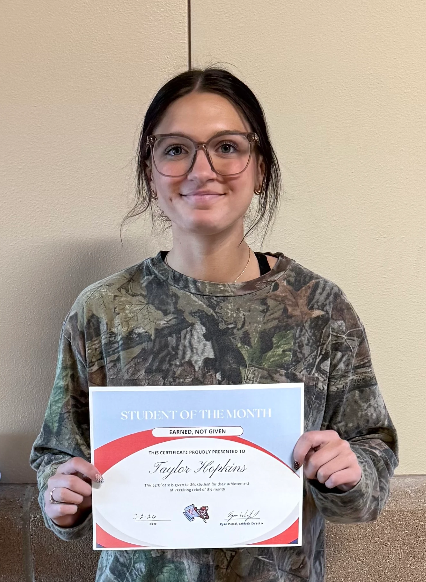 A young woman with glasses smiles while holding a "Student of the Month" certificate.