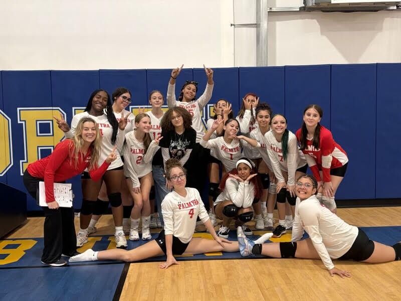 A volleyball team poses for a group photo in a gym with blue and yellow wall padding.