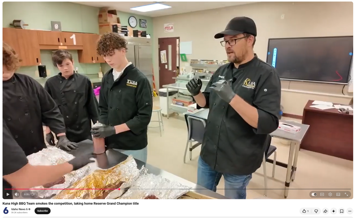 A chef in a black jacket and cap demonstrates grilling techniques to three students in a culinary arts kitchen.