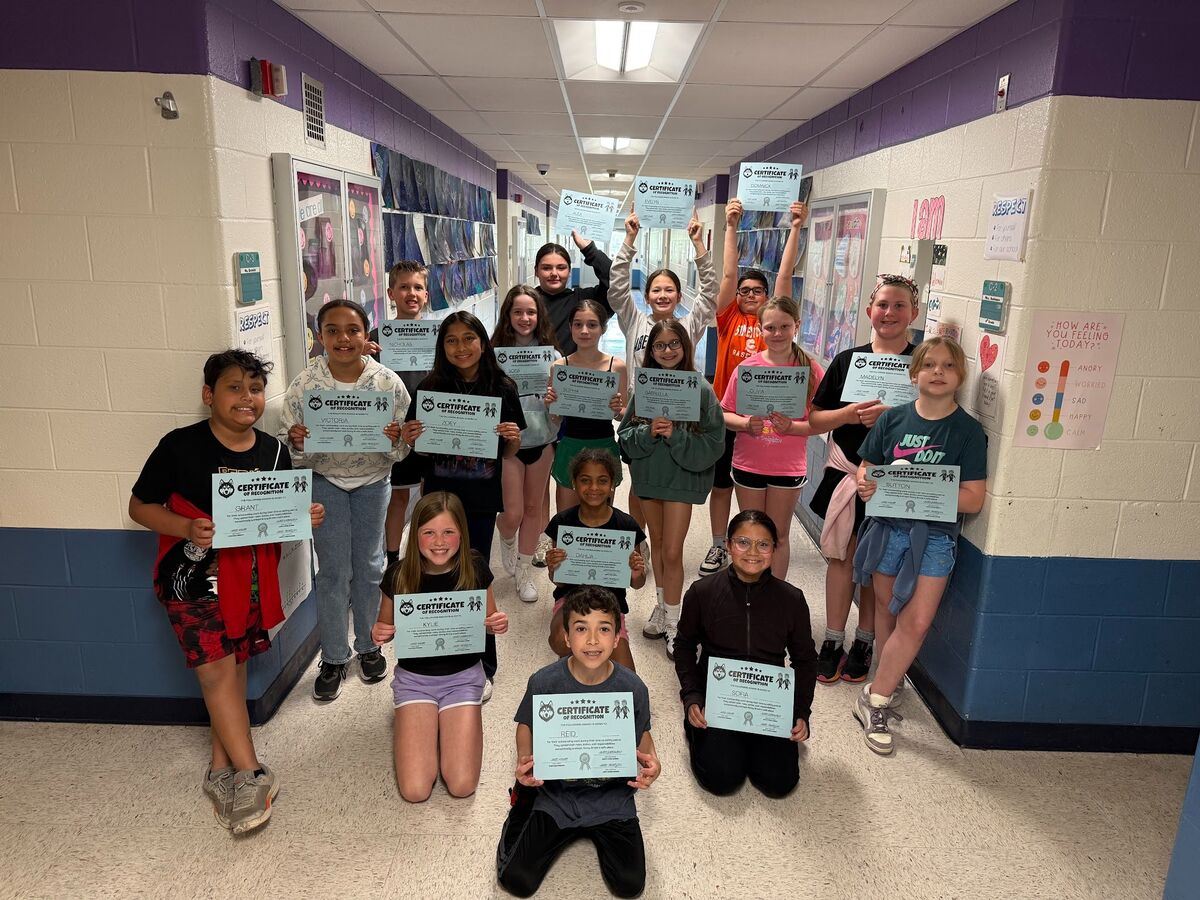 A diverse group of students proudly hold up certificates of recognition in a school hallway.